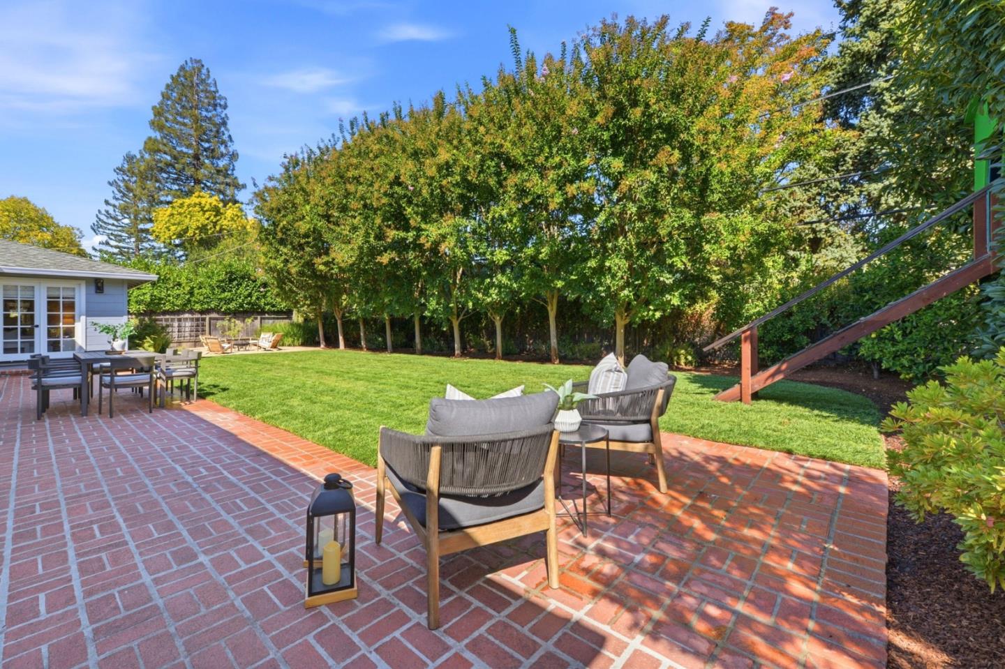 Backyard patio with red brick paving, outdoor seating, a broad lawn, and mature trees.