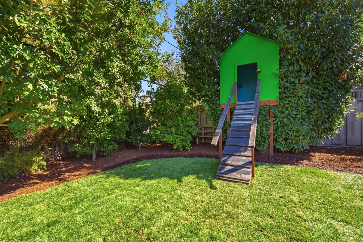 Backyard treehouse with a wooden ramp, leafy trees, and a sunny lawn.