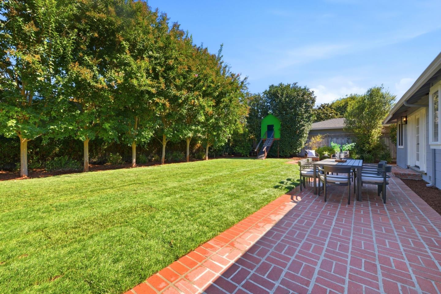 Backyard with a broad lawn, brick patio dining area, mature trees, and a green play structure.
