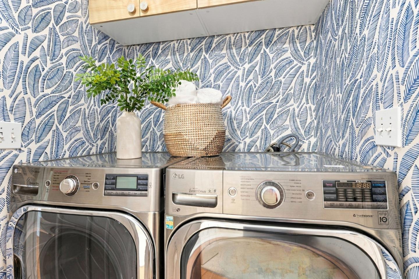 Laundry room with stainless steel washer and dryer, blue leaf-patterned wallpaper, and light wood cabinetry.