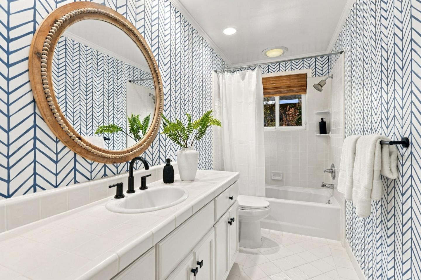 Bathroom with blue geometric wallpaper, a large round wood-framed mirror, and a white tub and vanity.