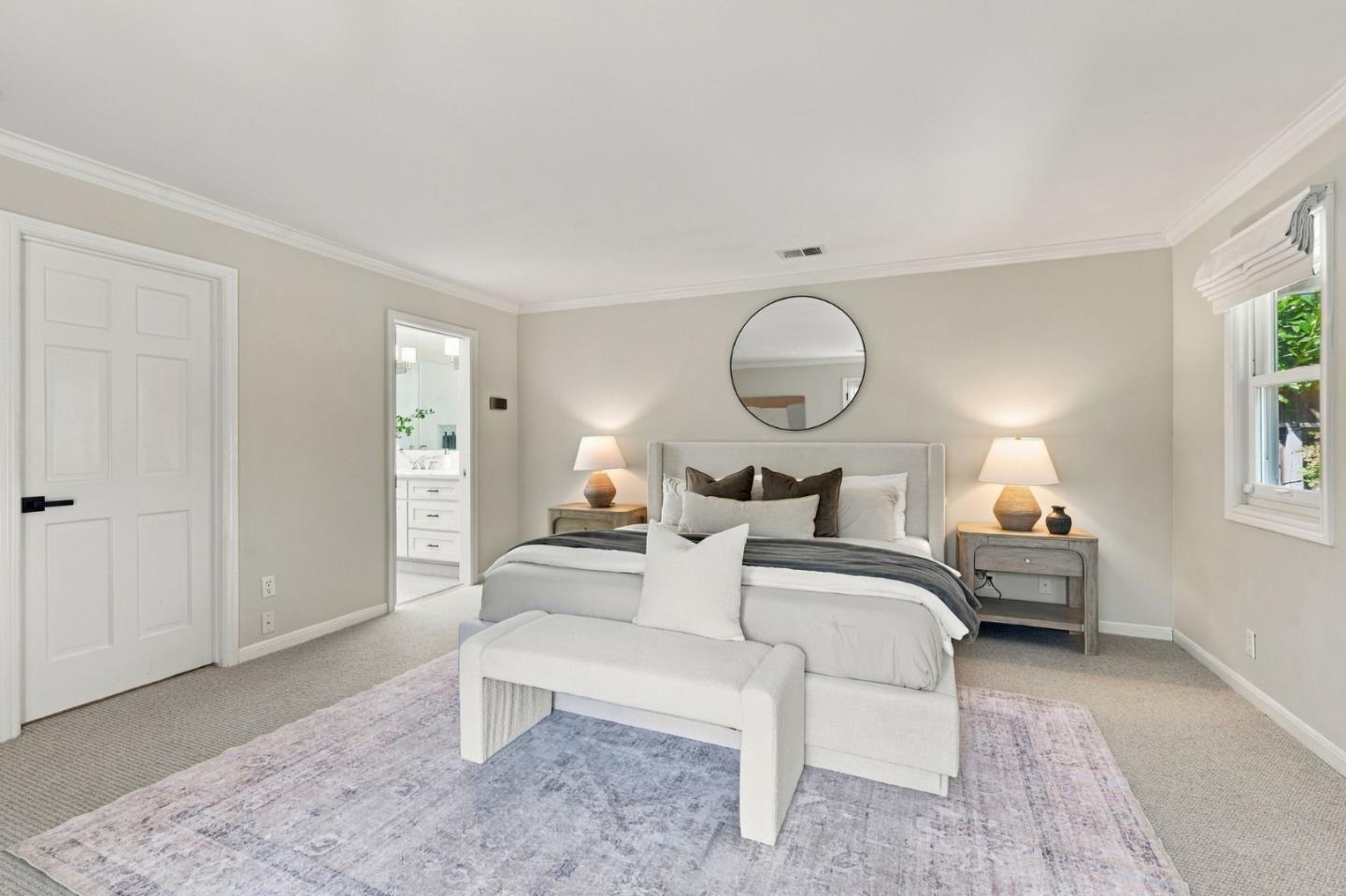 Staged bedroom with a white bed, round wall mirror, and doorway to a bright bathroom.