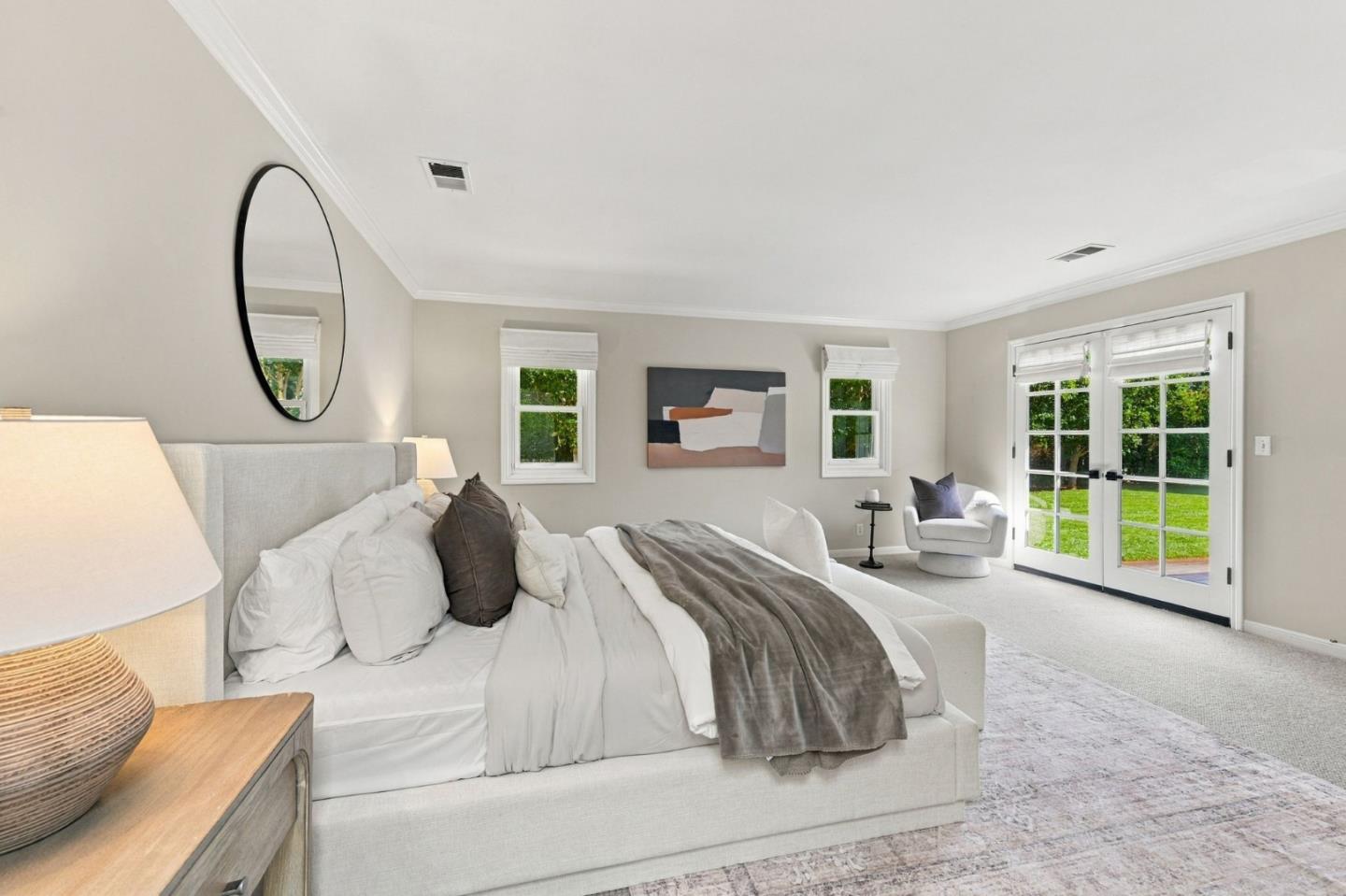 Staged bedroom with a large bed, round mirror, abstract art, and French doors overlooking the lawn.