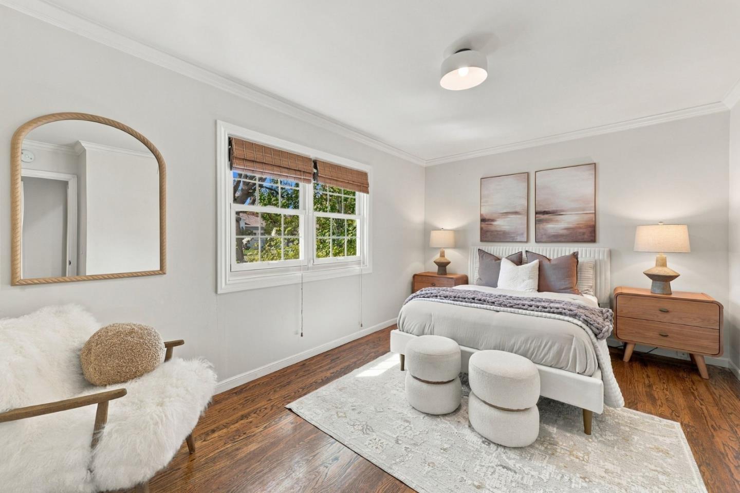 Staged bedroom with a white bed, wooden nightstands, abstract art, and a window with woven blinds.