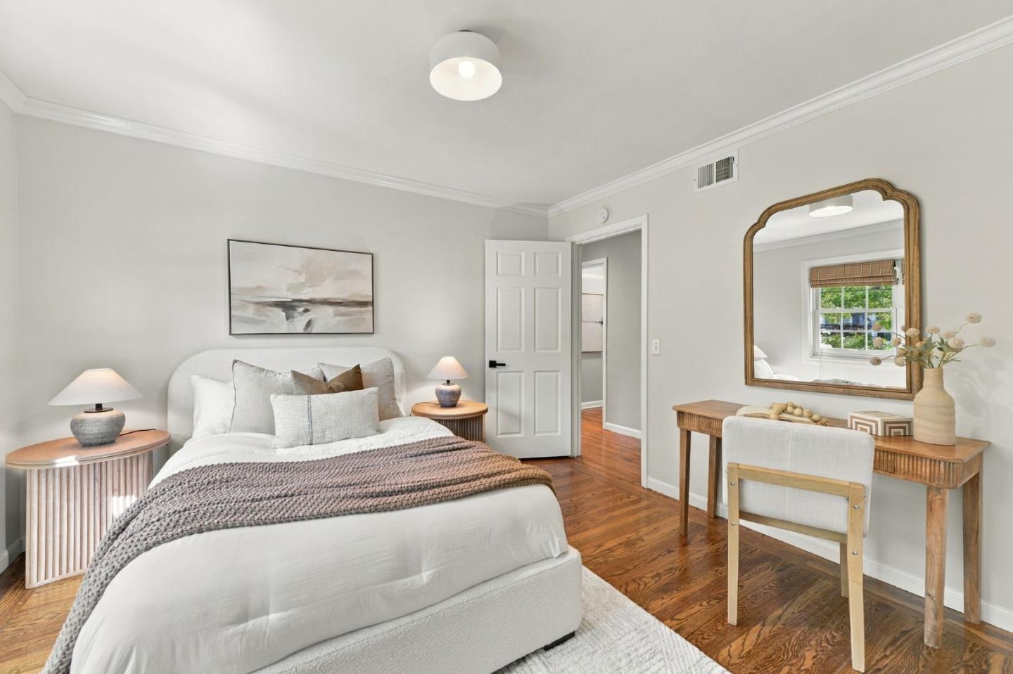 Staged bedroom with a white bed, neutral bedding, hardwood floors, and a desk with a large mirror.