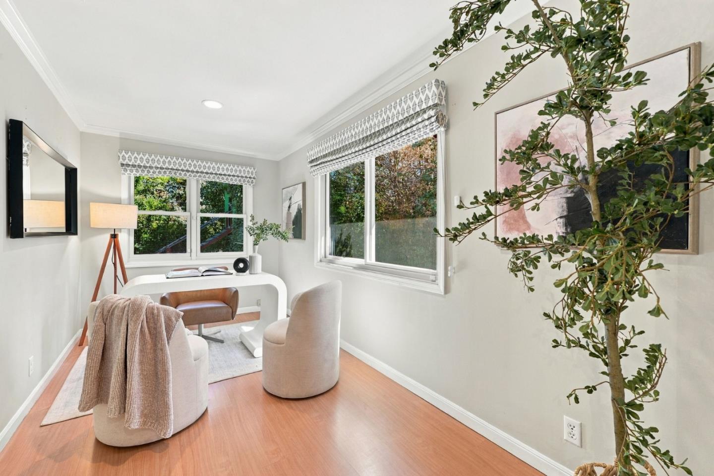 Staged home office with a white desk, large windows, patterned shades, and a leafy indoor tree.