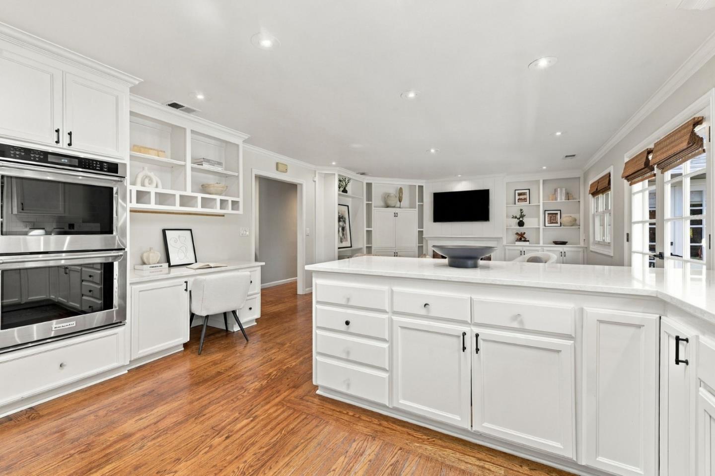 Bright white kitchen with a large island, built-in double ovens, hardwood floors, and wall shelving.