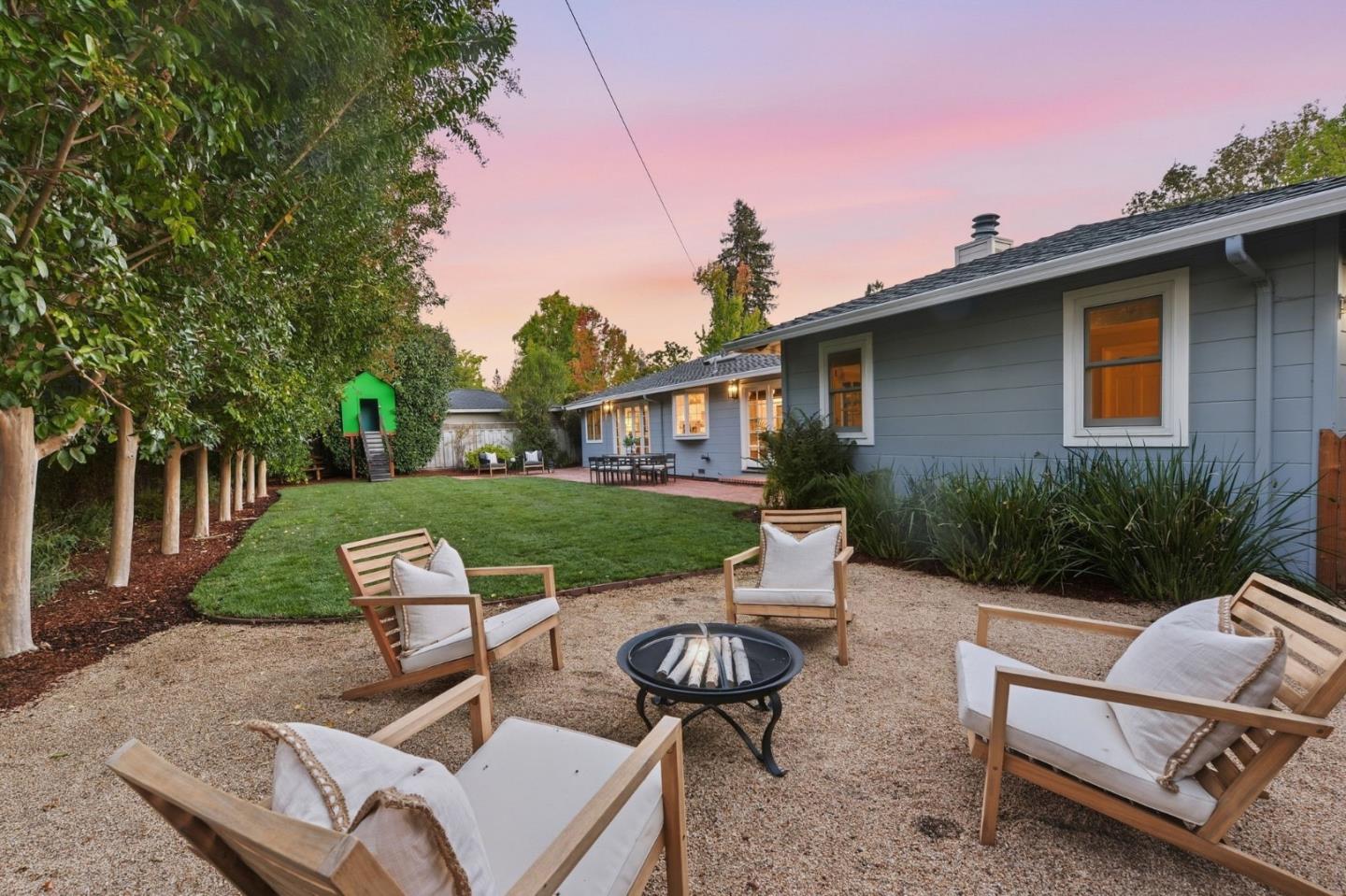 Backyard patio with cushioned chairs around a fire pit beside a gray house at dusk.