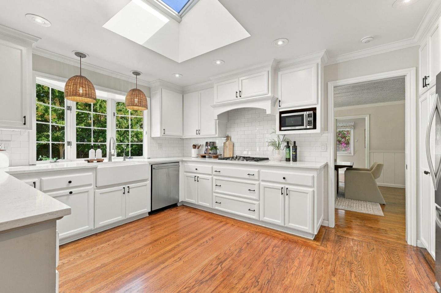 Bright white kitchen with a farmhouse sink beneath large windows, skylight, and warm hardwood floors.