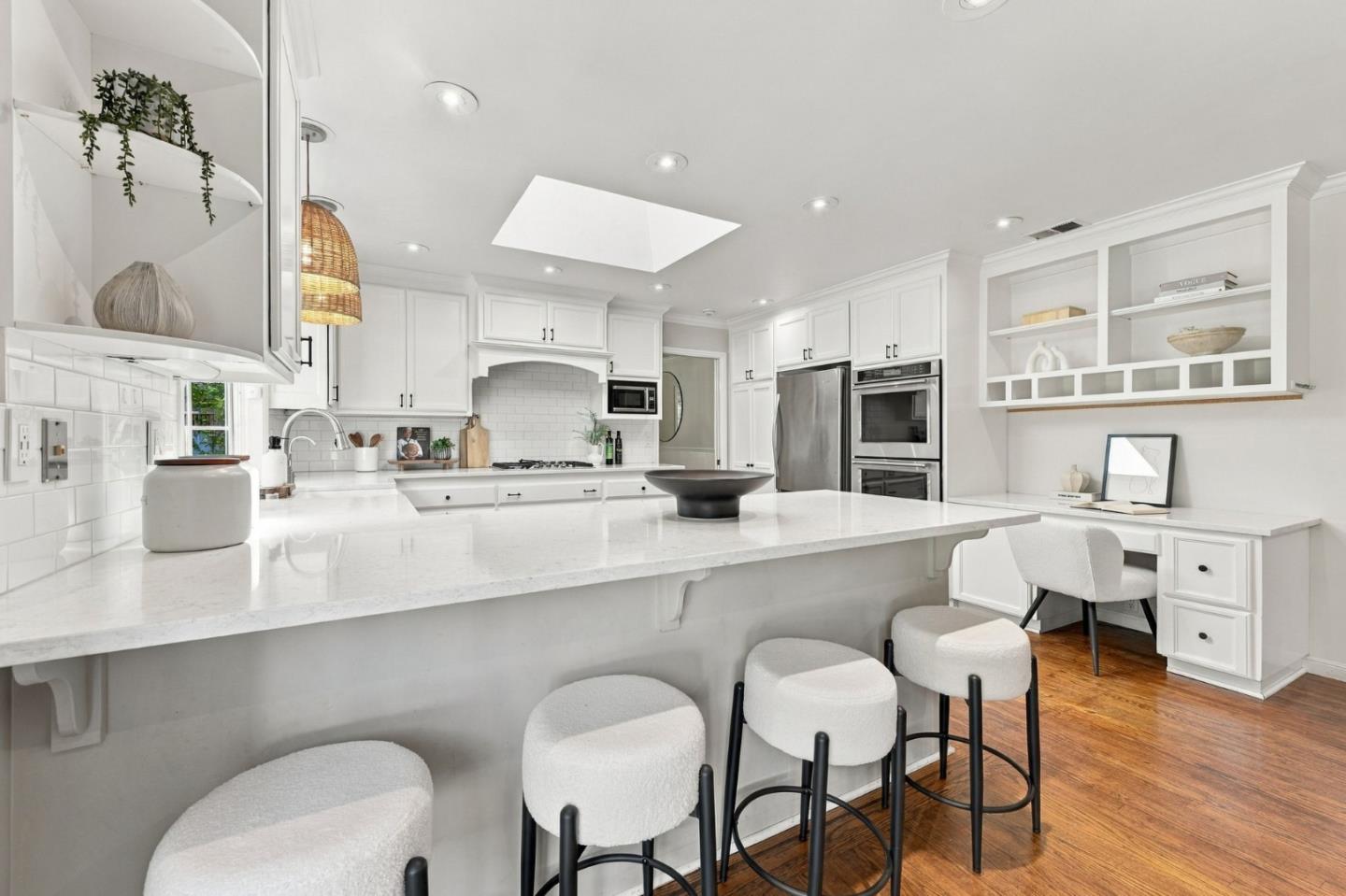 Bright white kitchen with a large island, upholstered stools, stainless steel appliances, and a skylight.