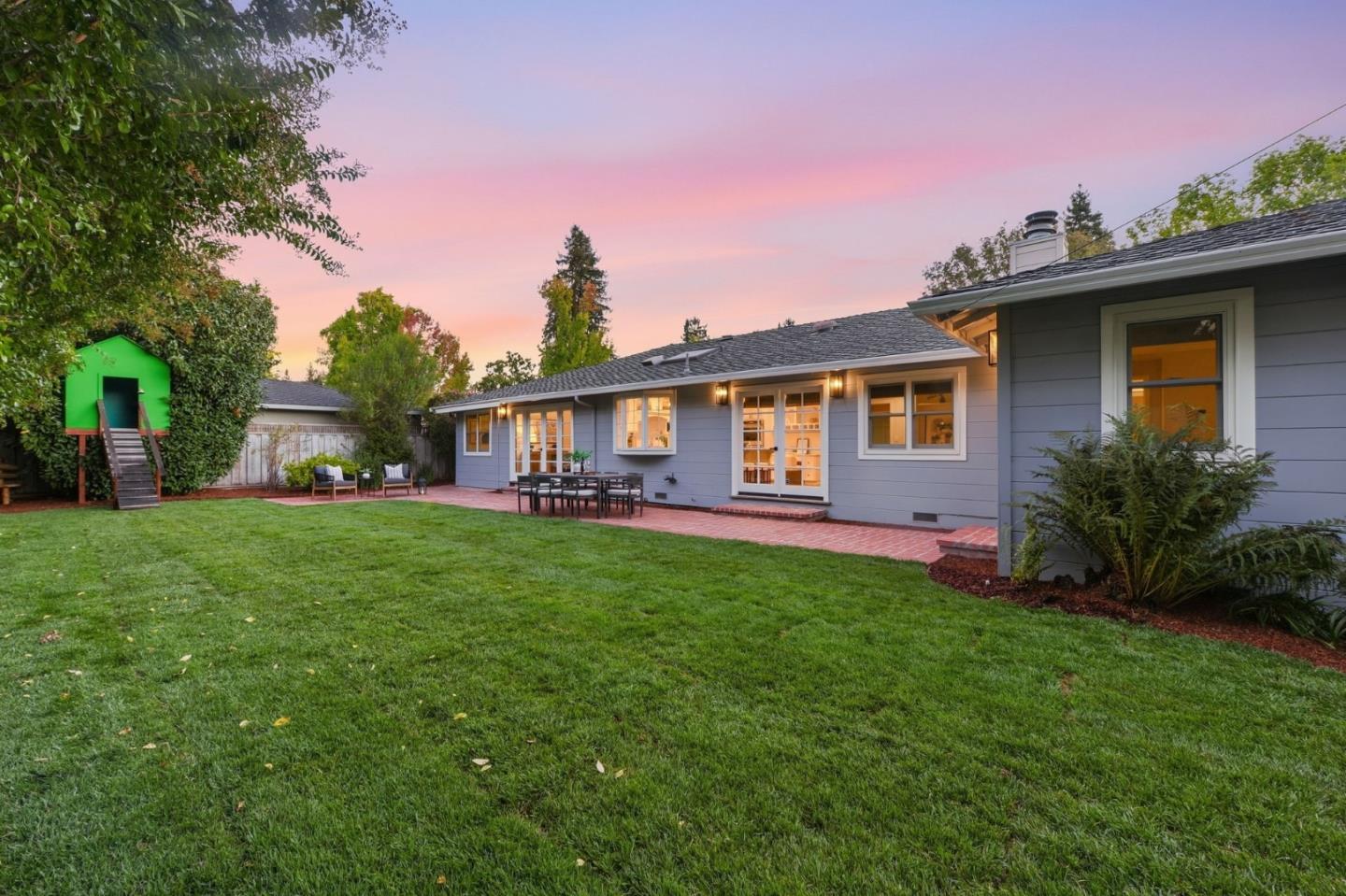 Gray single-story home behind a broad lawn, with a brick patio and green playhouse at dusk.