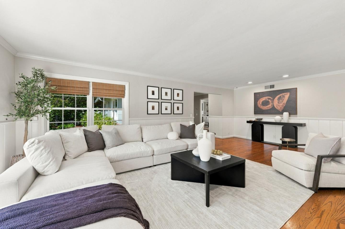 Staged living room with white sectional sofas, a black coffee table, and large windows with wood blinds.