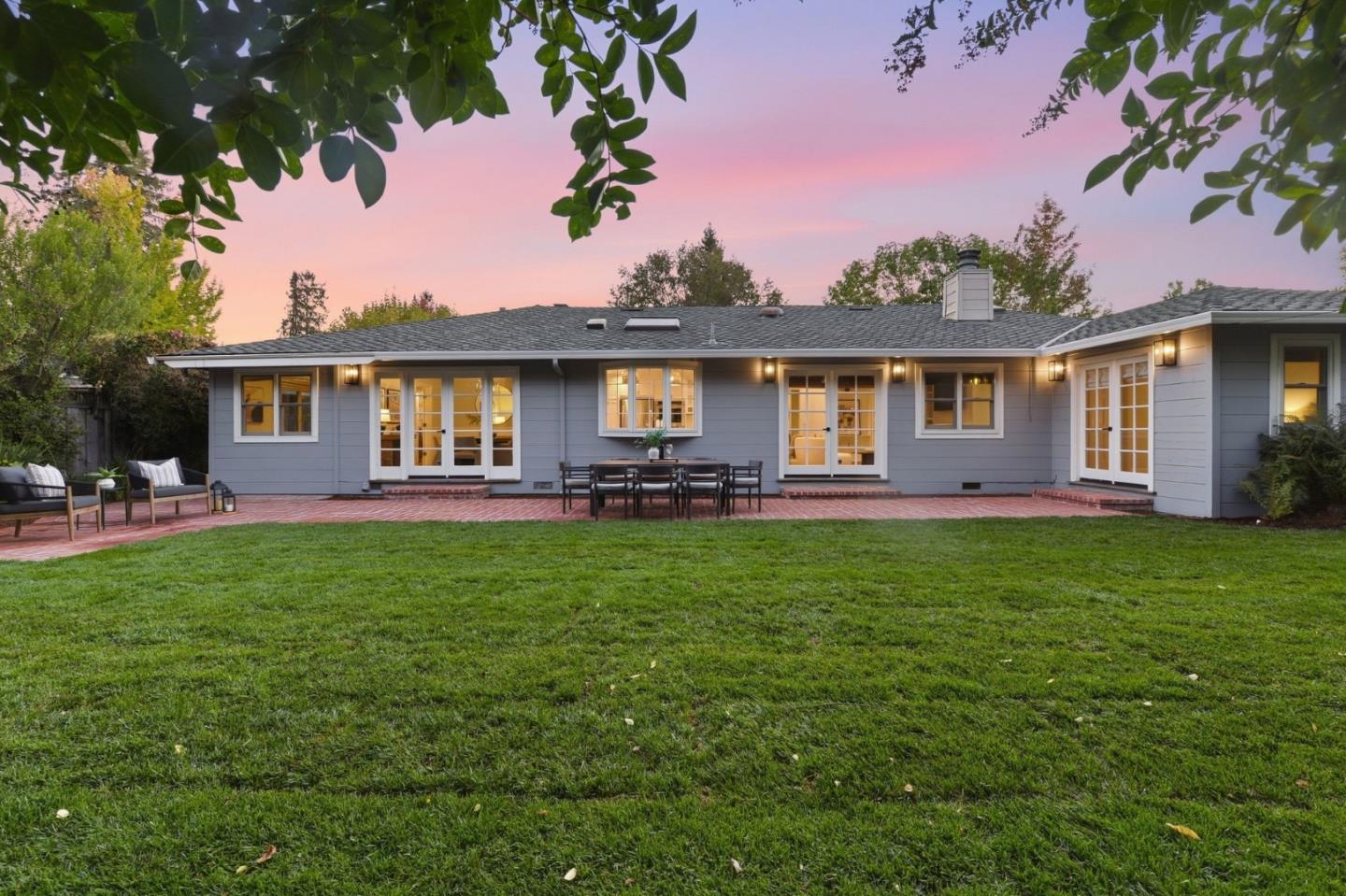 Rear of a gray home with French doors, brick patio, and lawn at dusk.