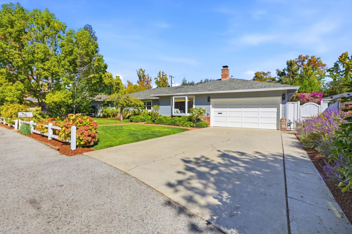 Single-story gray home with a white double garage, broad driveway, landscaped lawn, and white roadside fence.