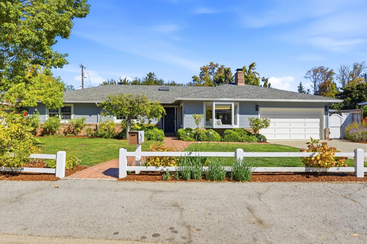 Single-story gray home with a white garage, landscaped lawn, brick walkway, and low white fence.