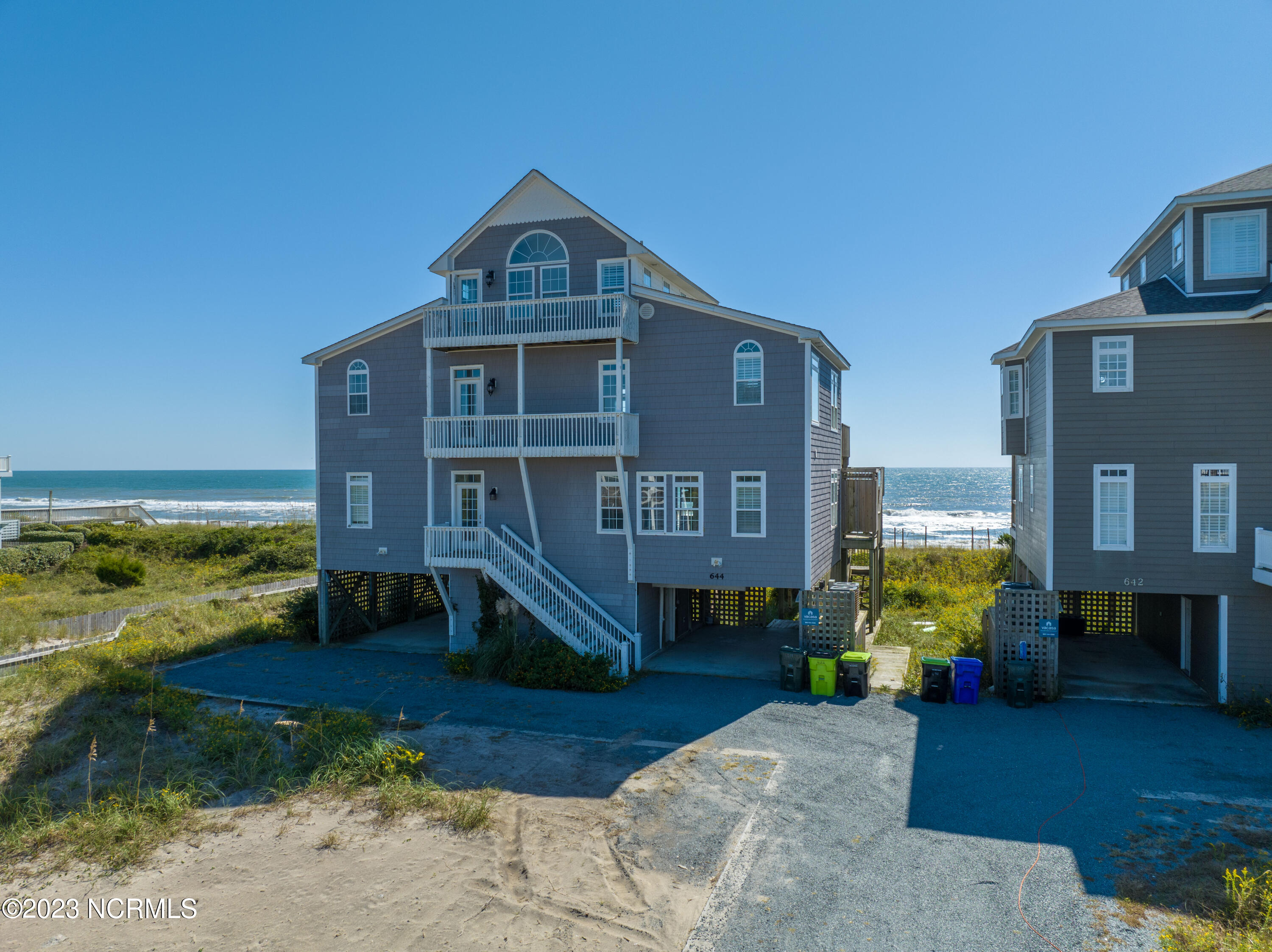 Gray multi-story beach house with white balconies beside the ocean and dune grass.