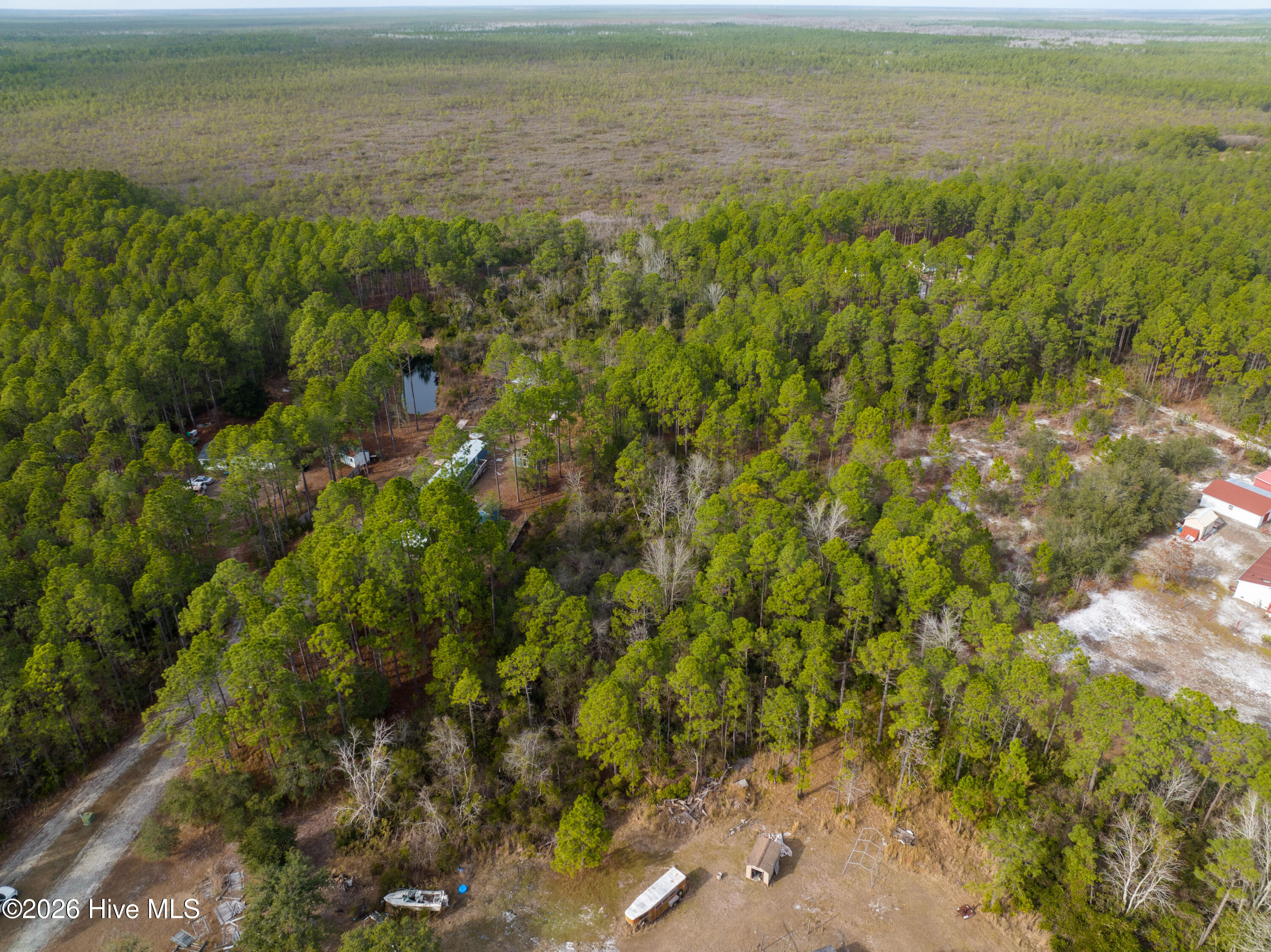 Aerial view of a wooded property with a pond, scattered buildings, and a cleared yard.