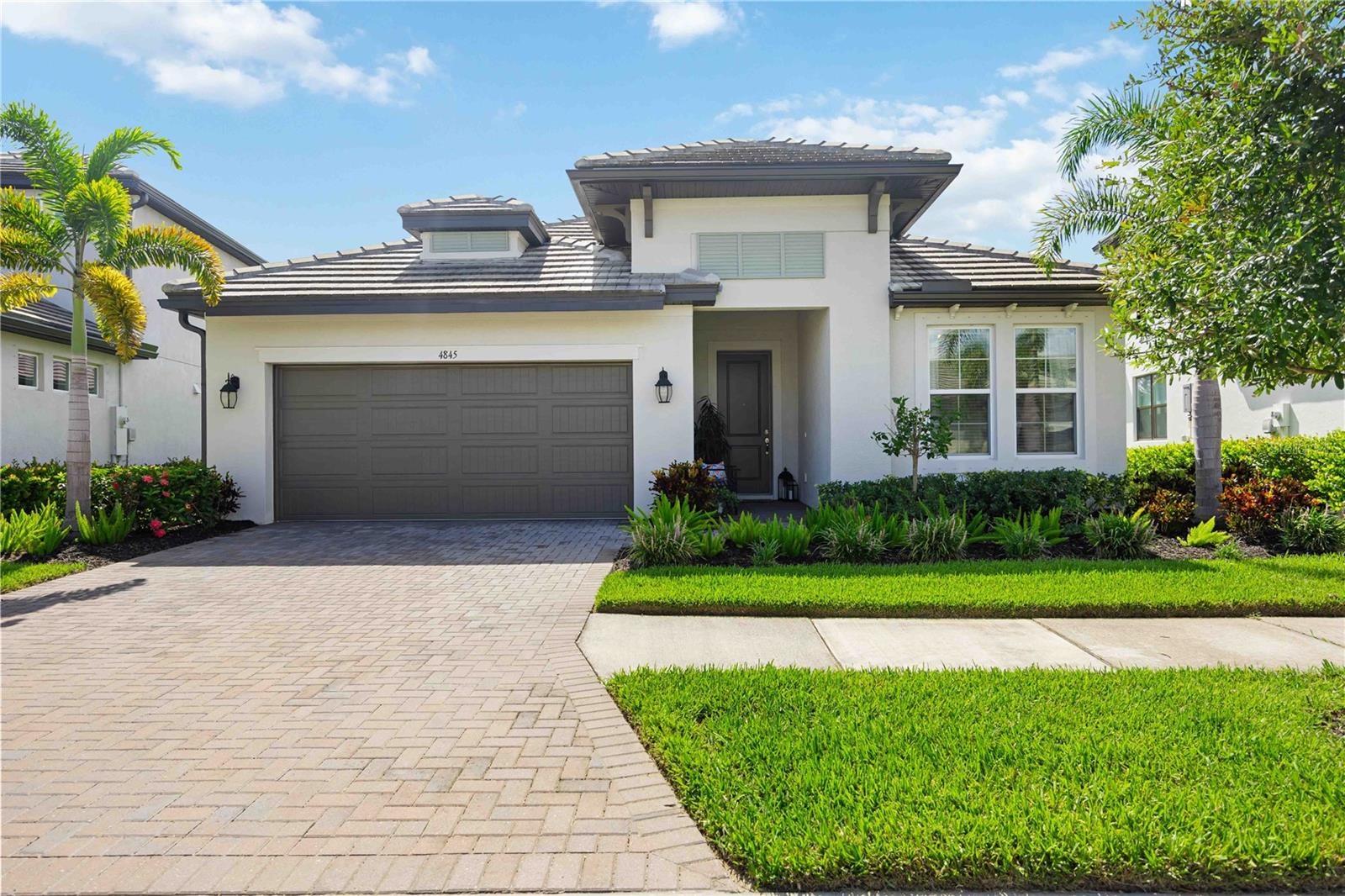 Modern white stucco home with a gray double garage door, paver driveway, and tropical landscaping.