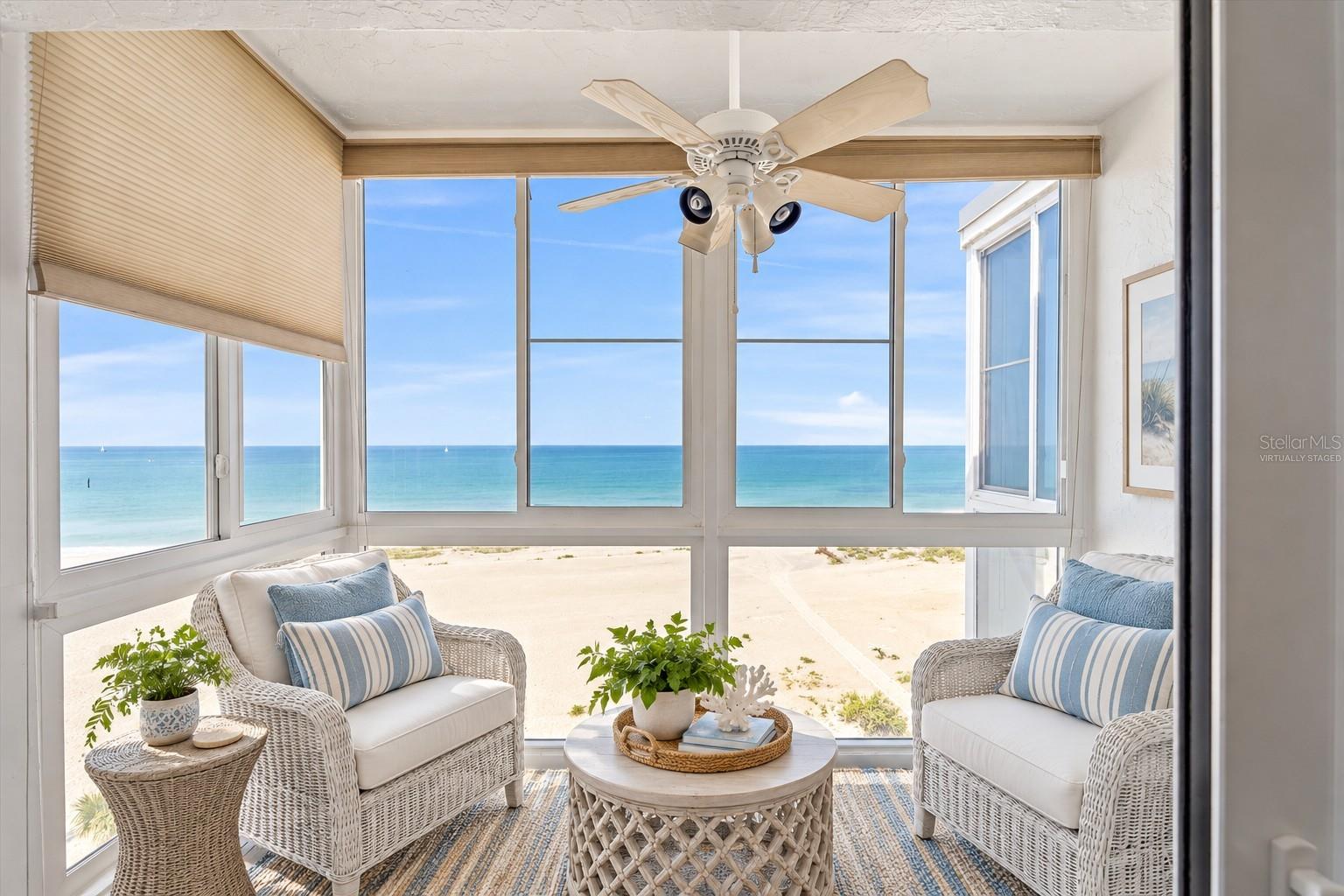 Enclosed sunroom with wicker seating and wraparound windows overlooking a sandy beach and turquoise water.