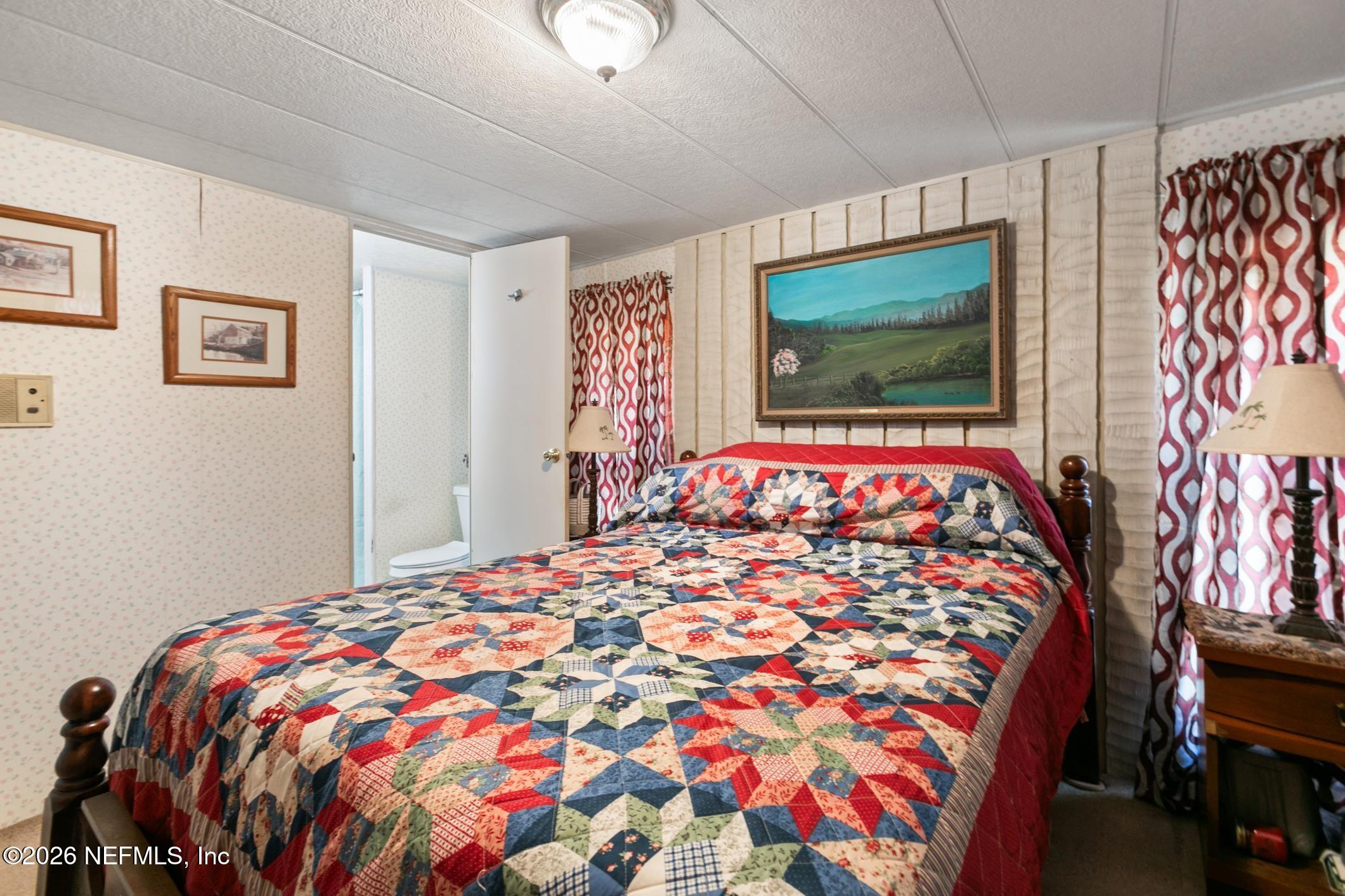 Bedroom with a colorful patchwork quilt, patterned curtains, and an open doorway to an adjoining bathroom.