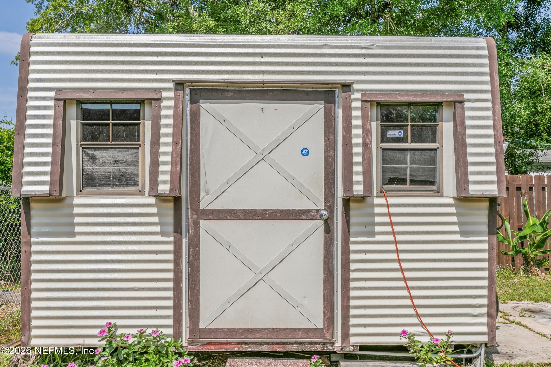 Small corrugated outbuilding with a cross-braced door, two windows, and flowering plants.