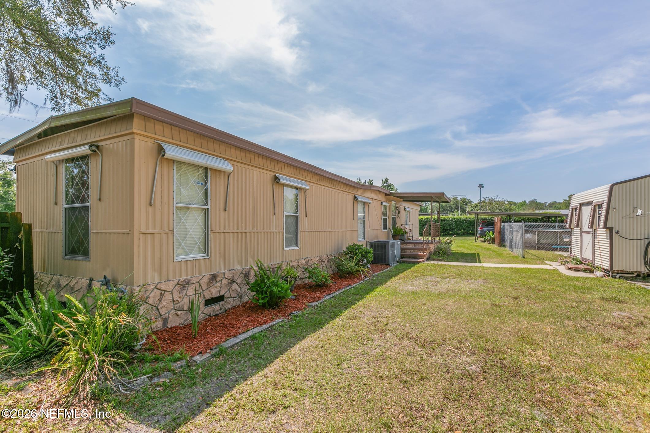 Tan manufactured home with a small deck, landscaped planting bed, and fenced grassy yard.