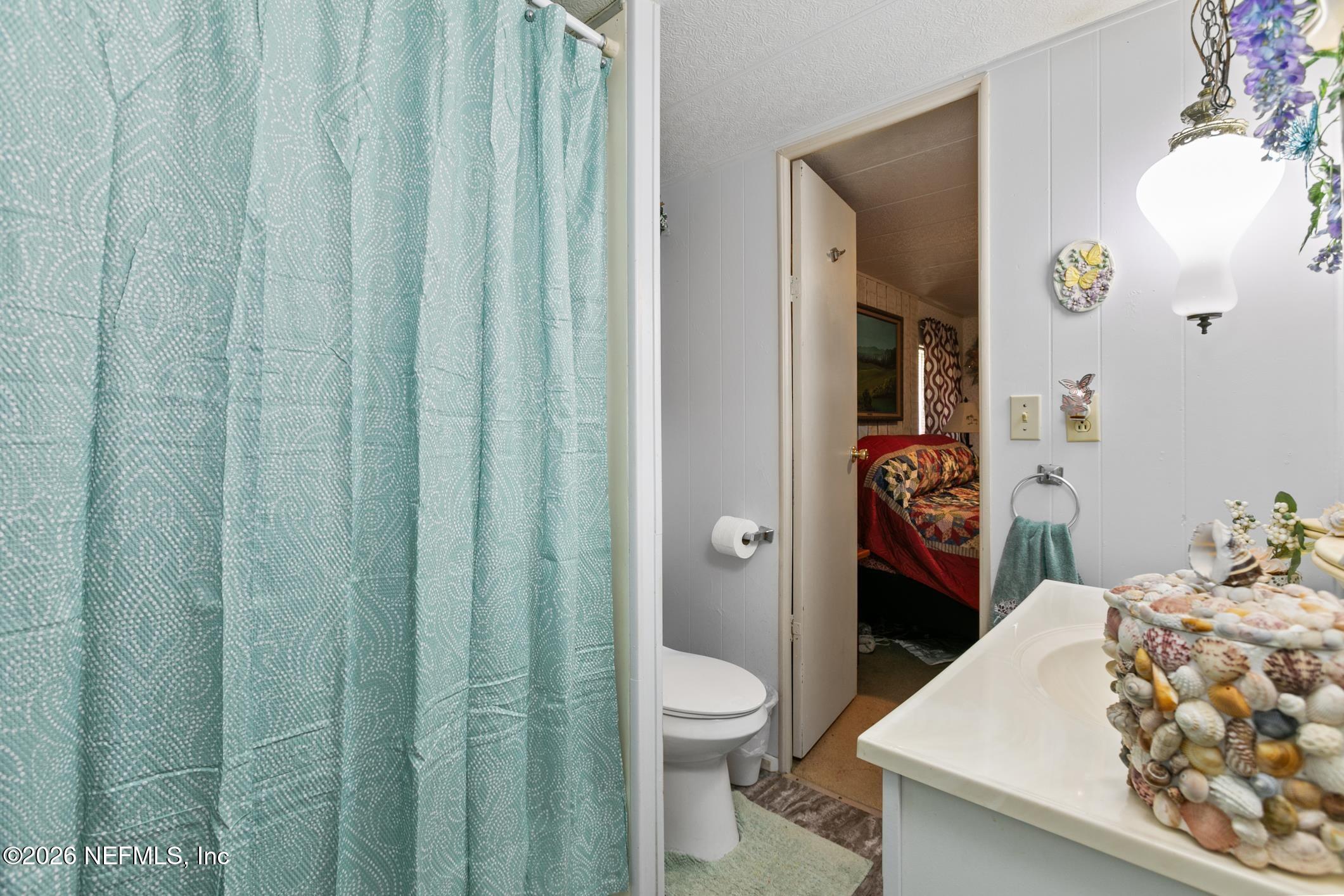 Bathroom with a turquoise shower curtain and seashell-decorated vanity, looking through an open doorway to a bedroom.