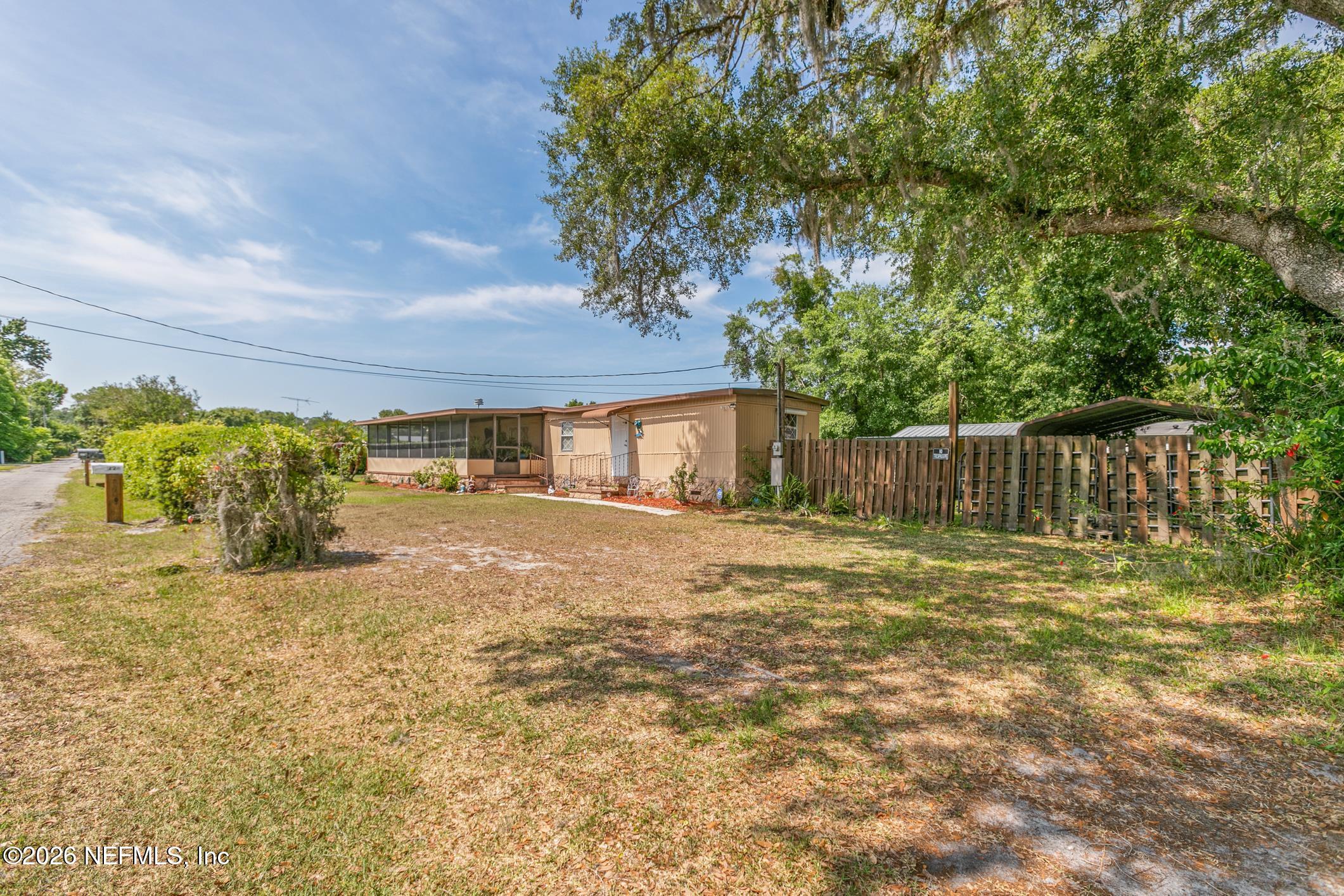 Tan manufactured home with a screened porch, fenced carport, and broad grassy yard beneath a large oak tree.