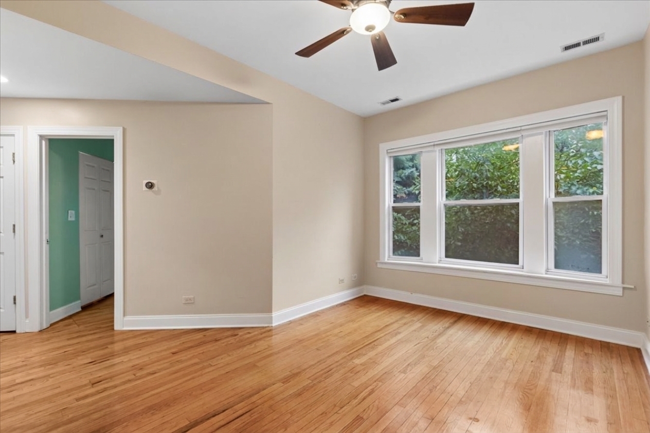 Vacant room with hardwood floors, ceiling fan, and wide windows overlooking greenery.