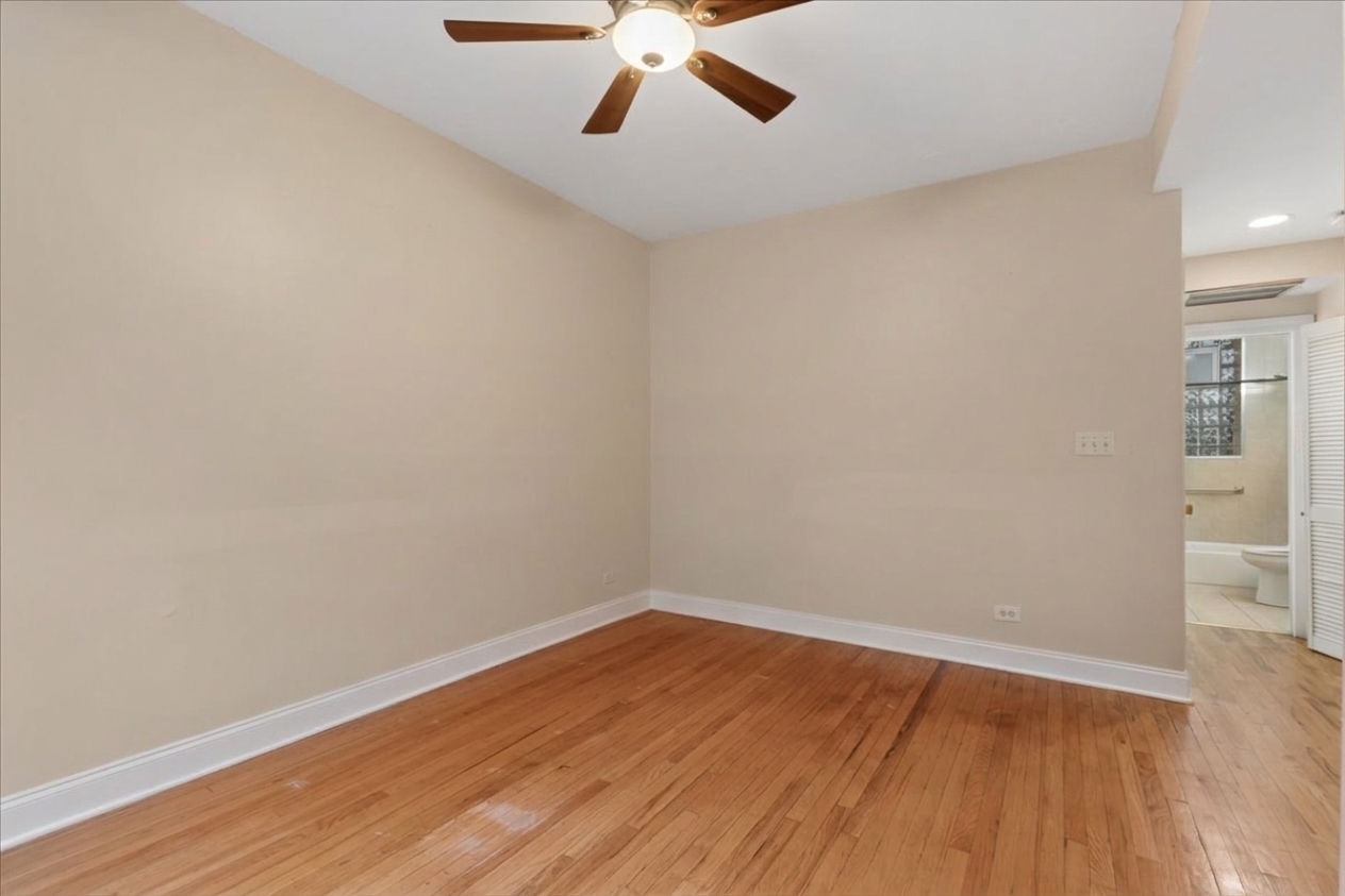 Empty bedroom with hardwood floors and a ceiling fan, with a bathroom visible through the doorway.