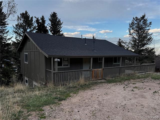 Dark gray house with a long covered porch beside a gravel driveway and evergreen trees.