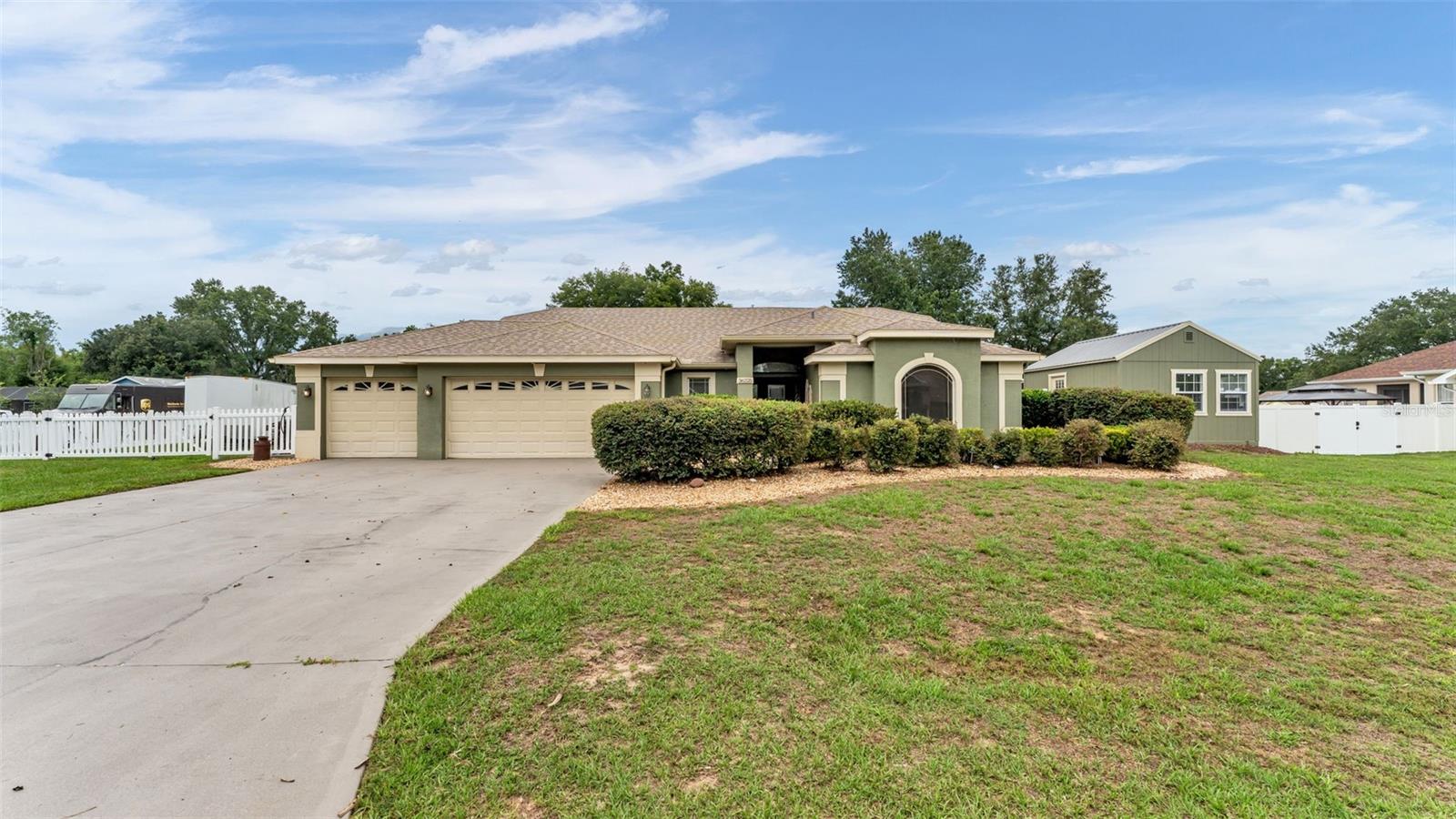 Single-story green home with a three-car garage, broad driveway, front hedges, and spacious lawn.