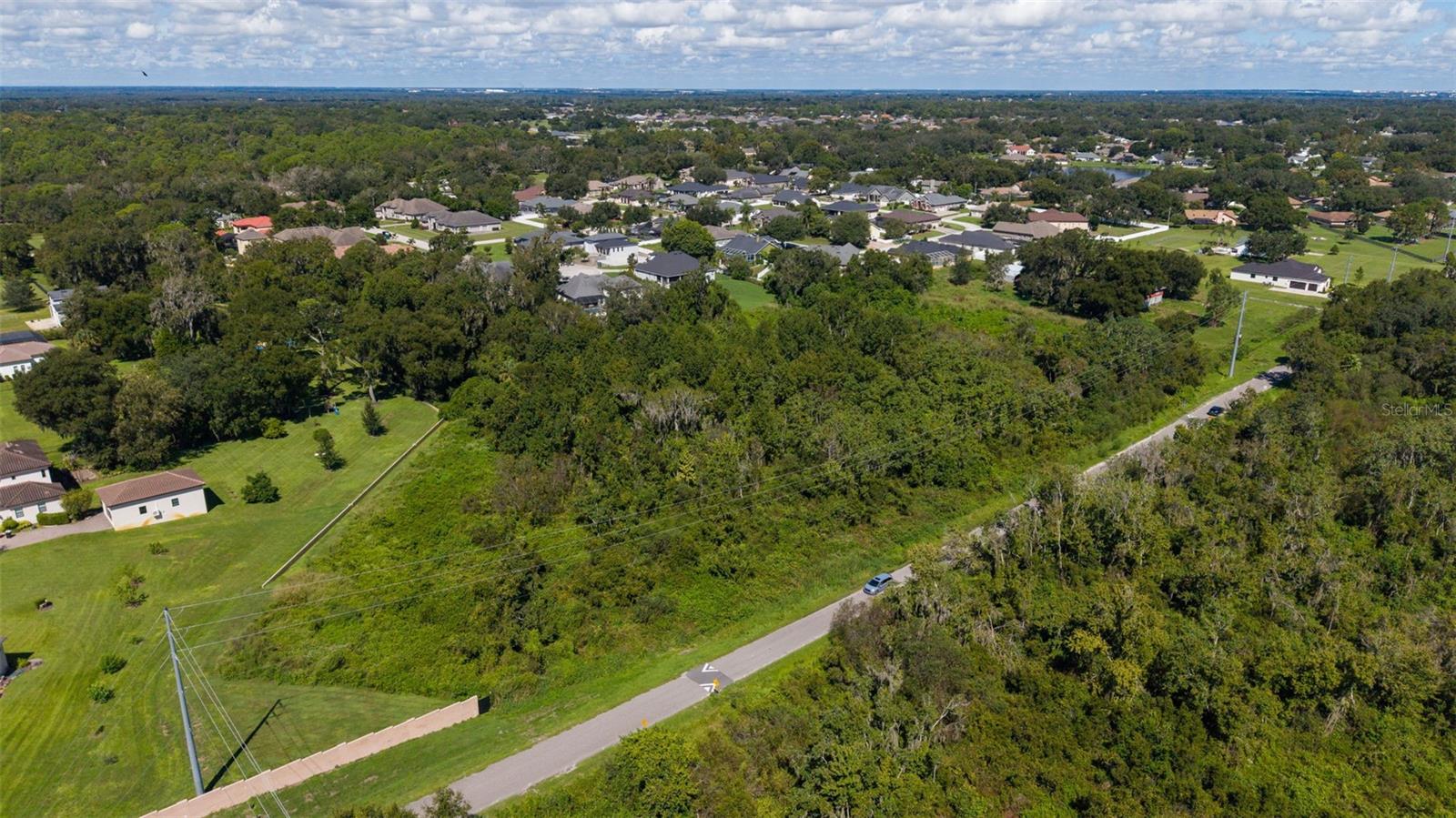 Aerial view of wooded vacant land beside a road, with homes and grassy lots nearby.