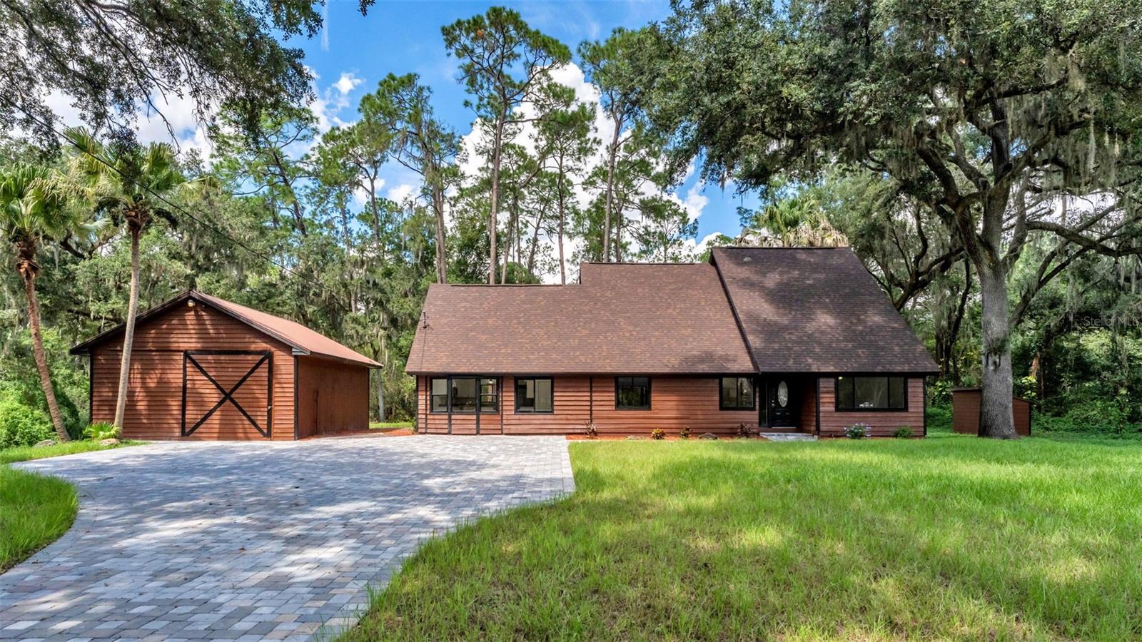 Wood-sided home with a steep roof, matching outbuilding, paved driveway, and mature trees.