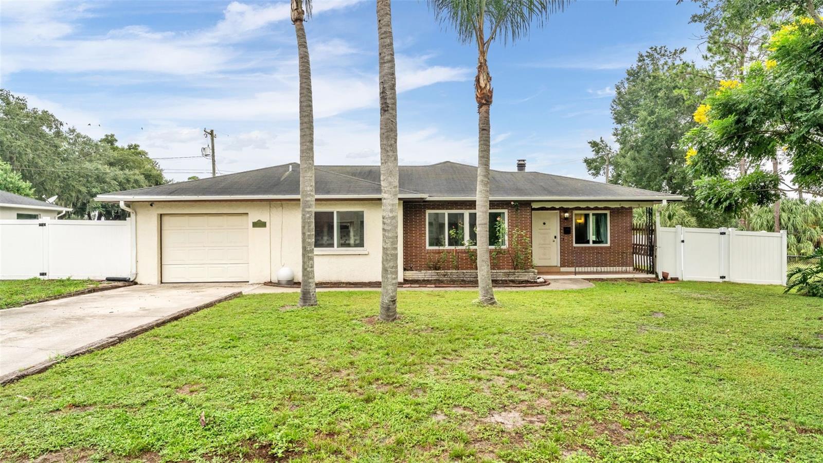 Single-story home with a brick-and-stucco facade, attached garage, palm trees, and a white fenced yard.