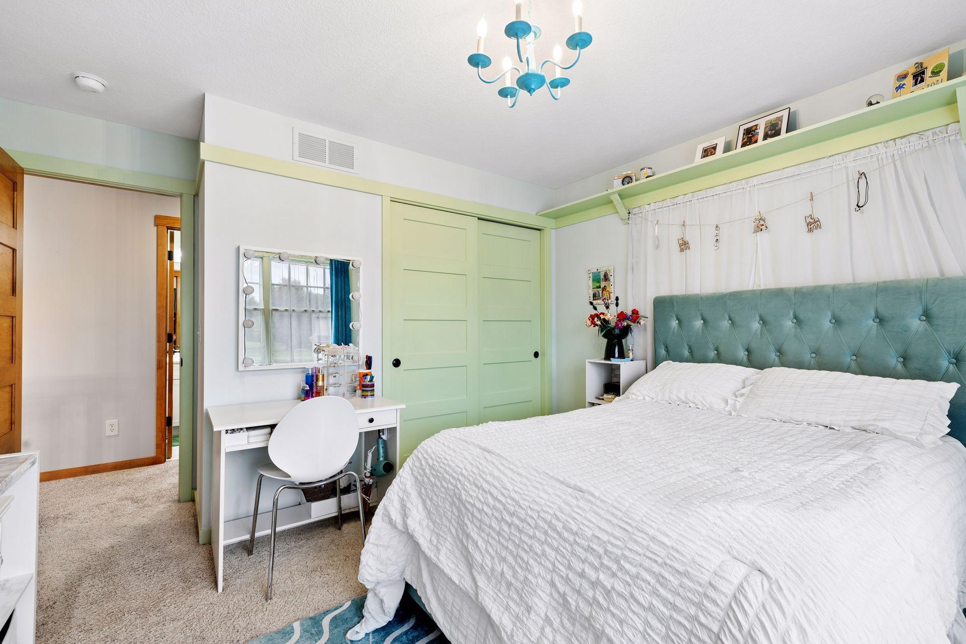 Bedroom with a white bed, teal tufted headboard, pale green closet doors, and a small vanity desk.