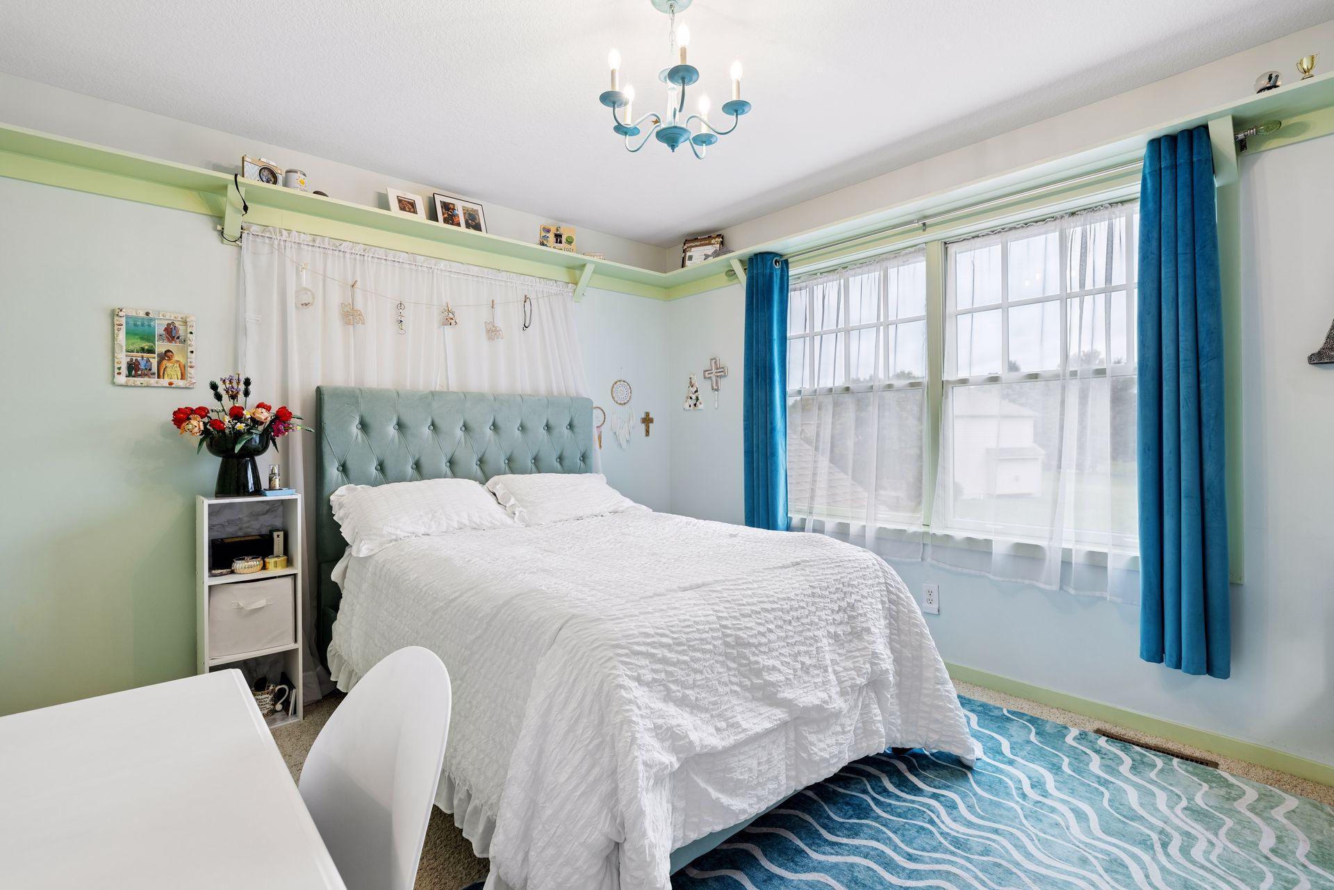 Bedroom with a white bed, aqua tufted headboard, blue curtains, and large windows.
