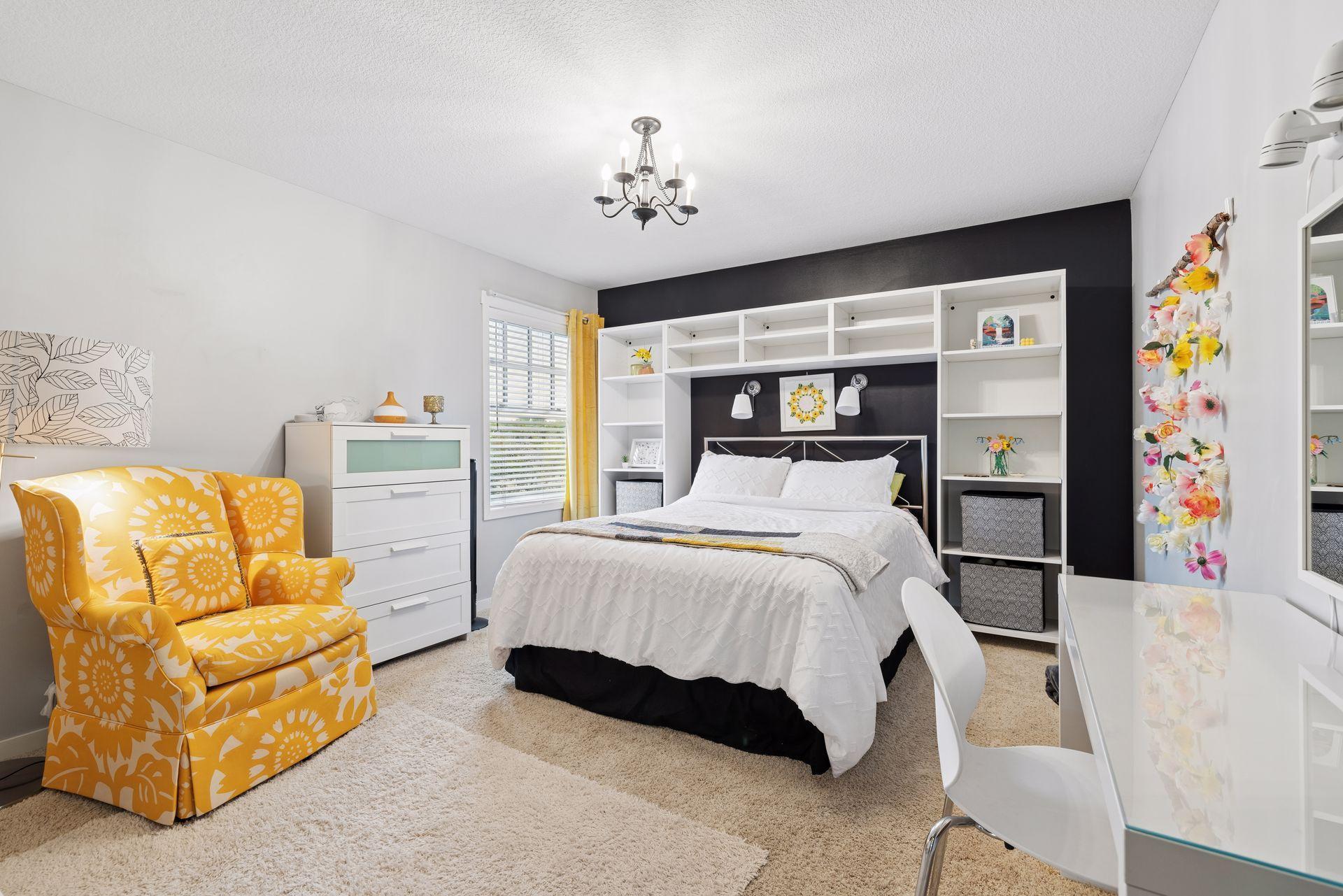 Bedroom with a white bed, built-in shelving, dark accent wall, and yellow floral armchair.