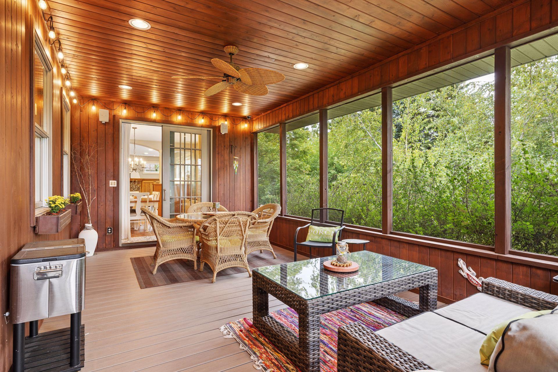 Enclosed wood-paneled porch with wicker seating and large windows overlooking leafy trees.