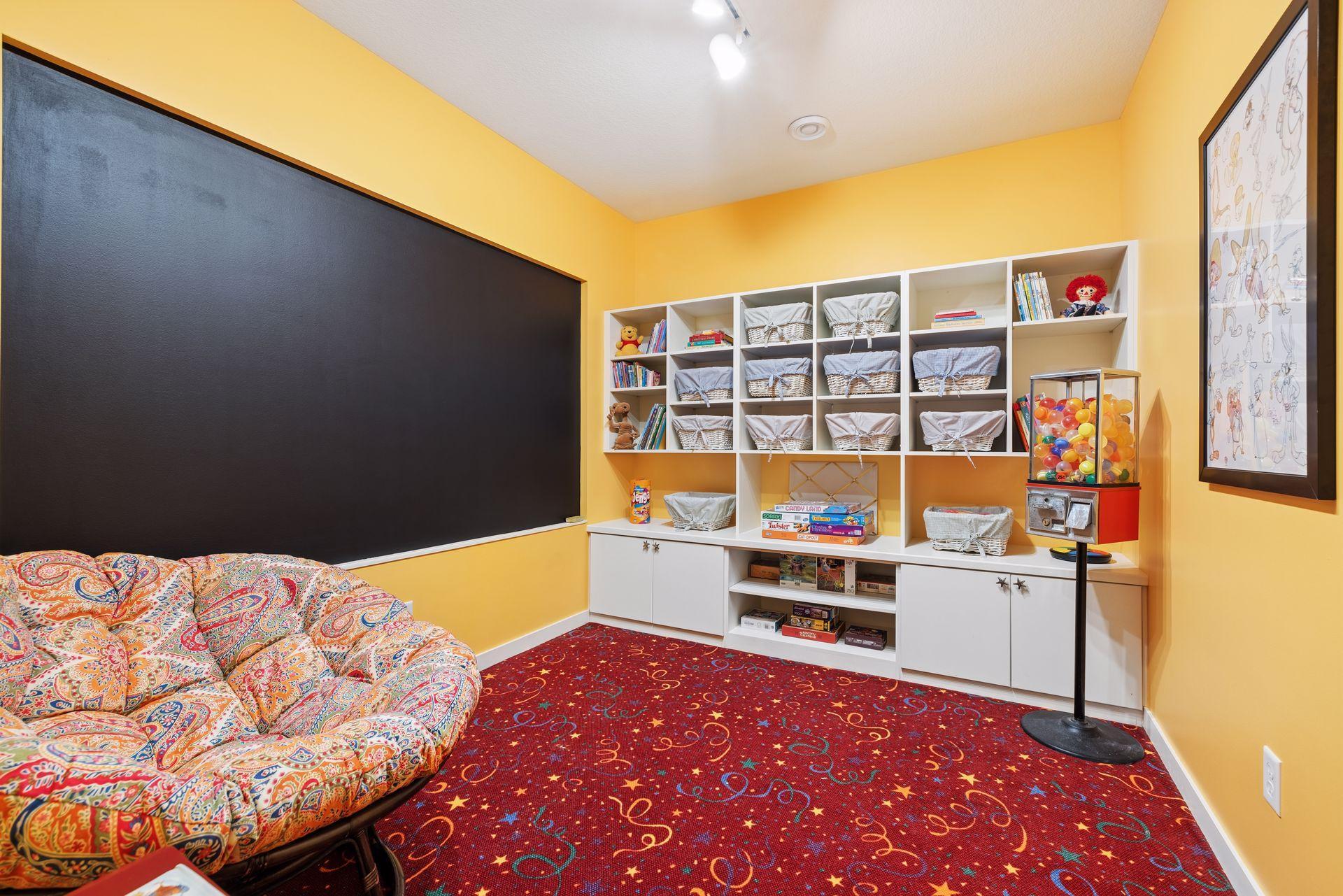 Playroom with a large chalkboard, toy-filled built-in shelves, patterned sofa, and colorful carpet.