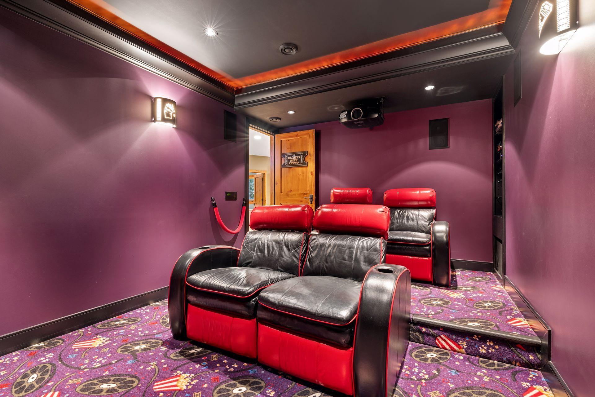 Home theater with red-and-black leather recliners, purple walls, and patterned movie-themed carpet.
