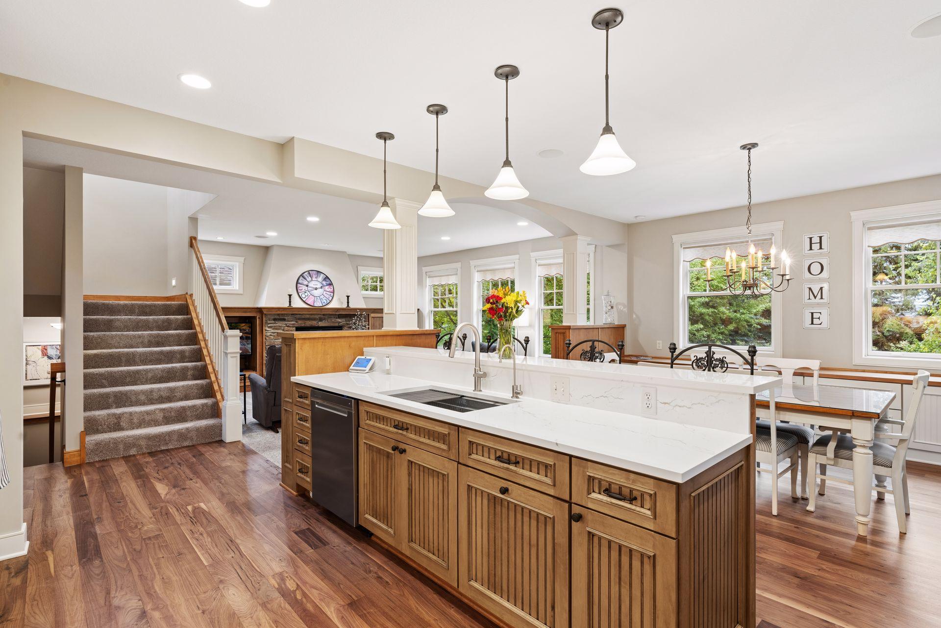 Open kitchen with a white island and wood cabinetry beside a dining area, staircase, and stone fireplace.