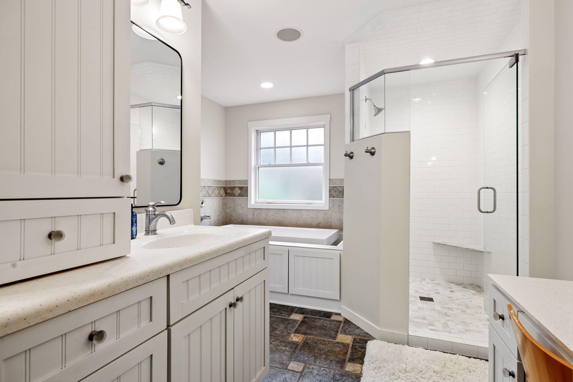 Bathroom with a glass subway-tile shower, bathtub beneath a window, and white paneled vanity.