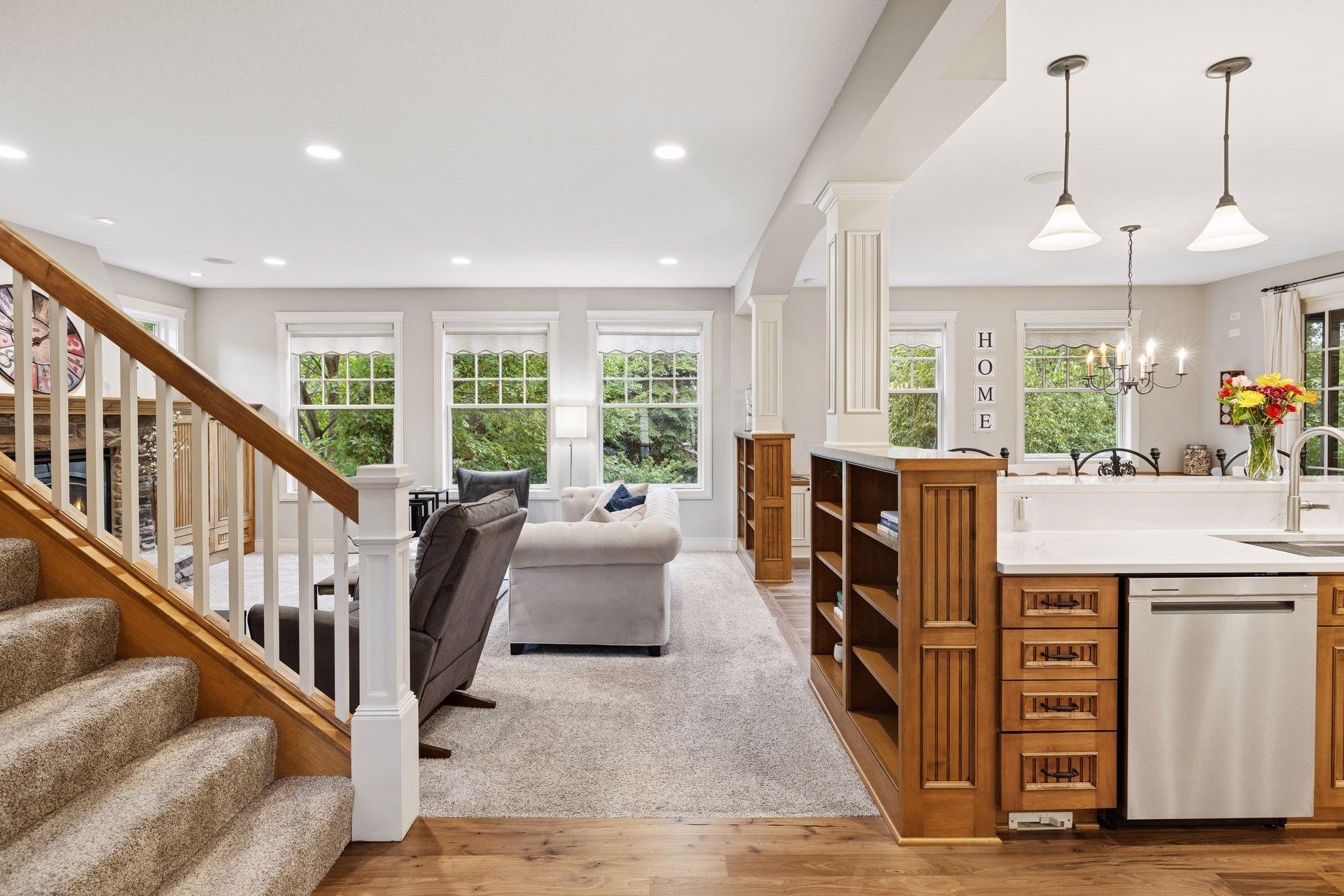 Open living room and kitchen with carpeted staircase, wood cabinetry, and large windows overlooking greenery.