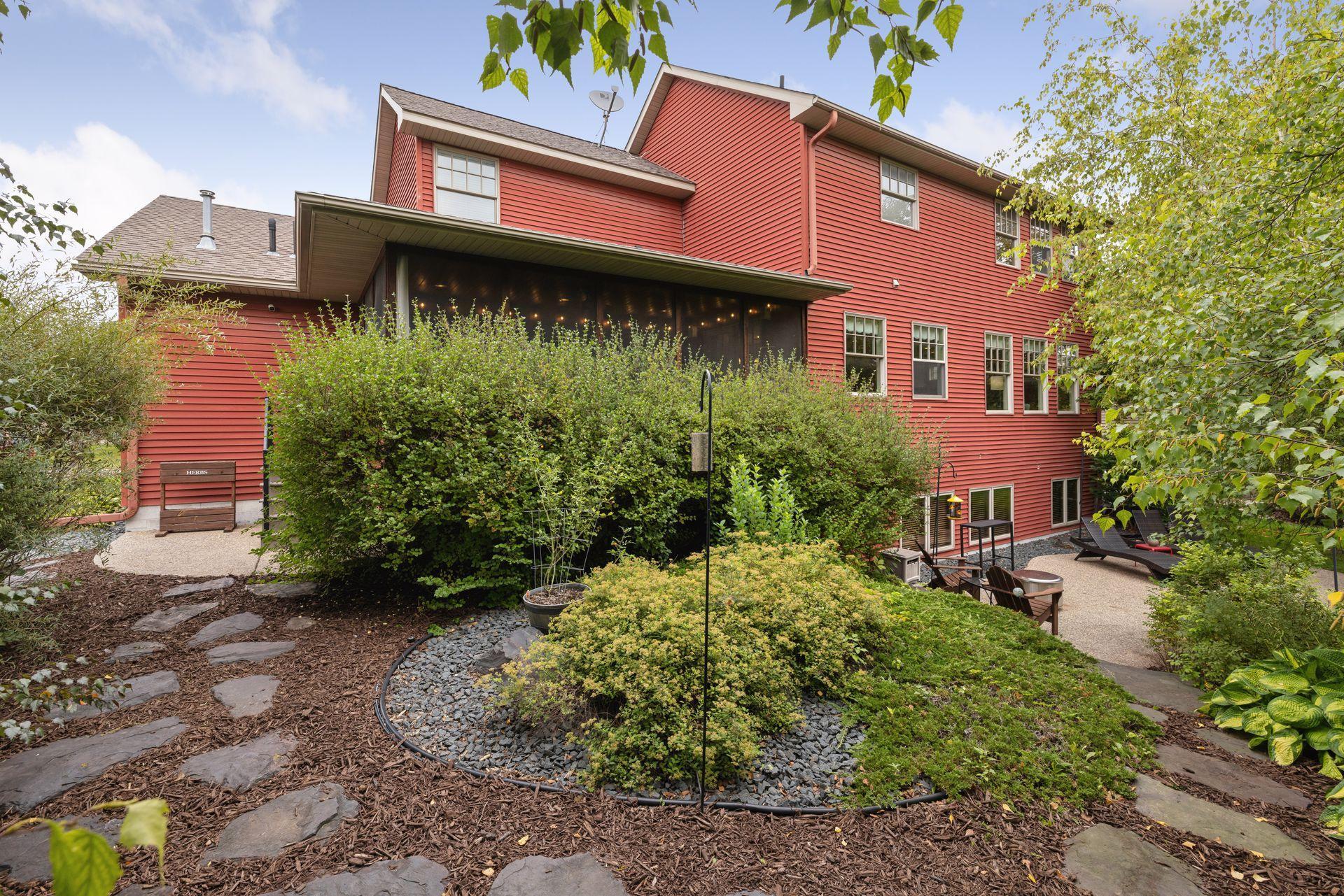 Red two-story house viewed from a landscaped backyard with stone paths and a covered patio.