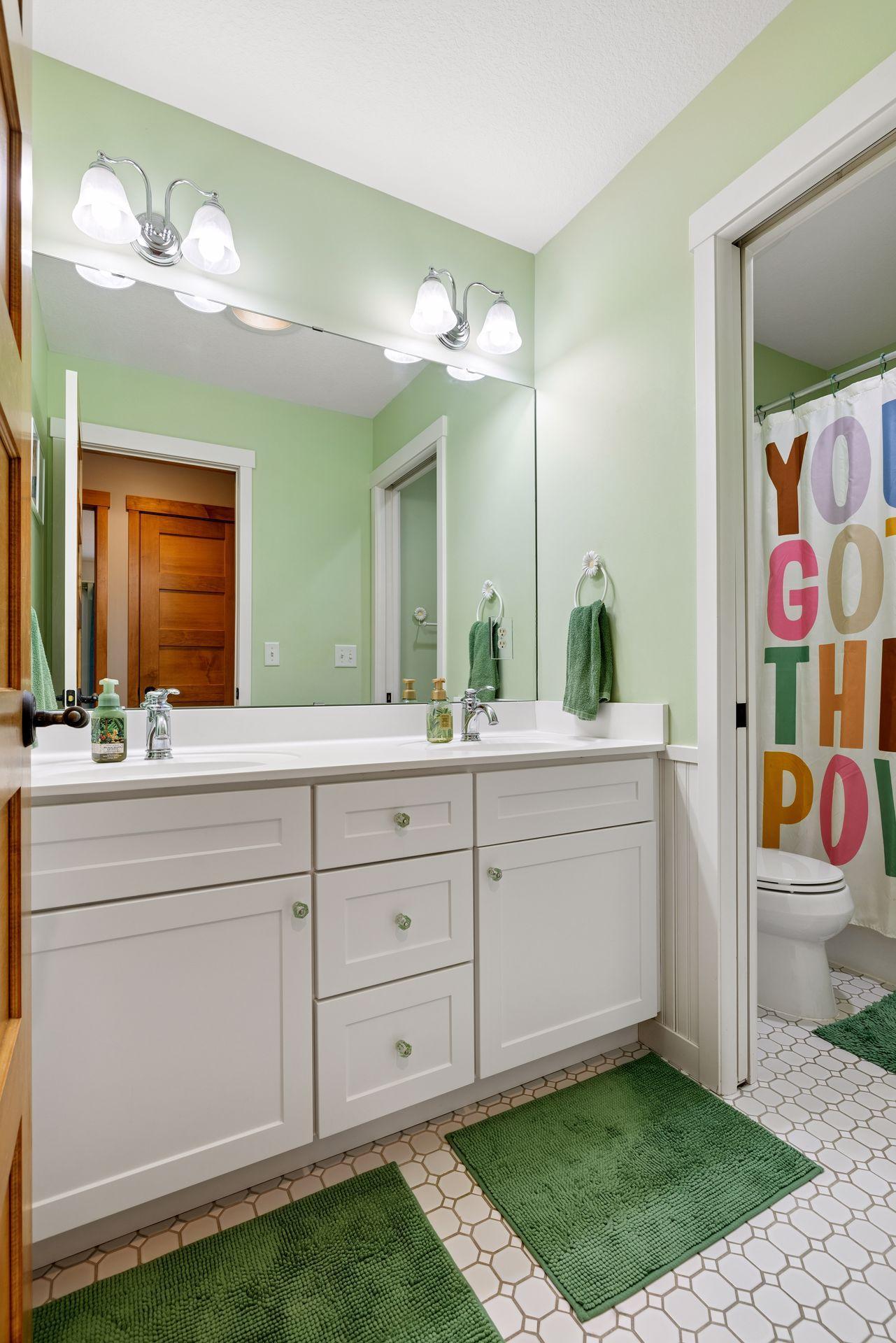 Bathroom with a white double-sink vanity, large mirror, pale green walls, and green bath mats.