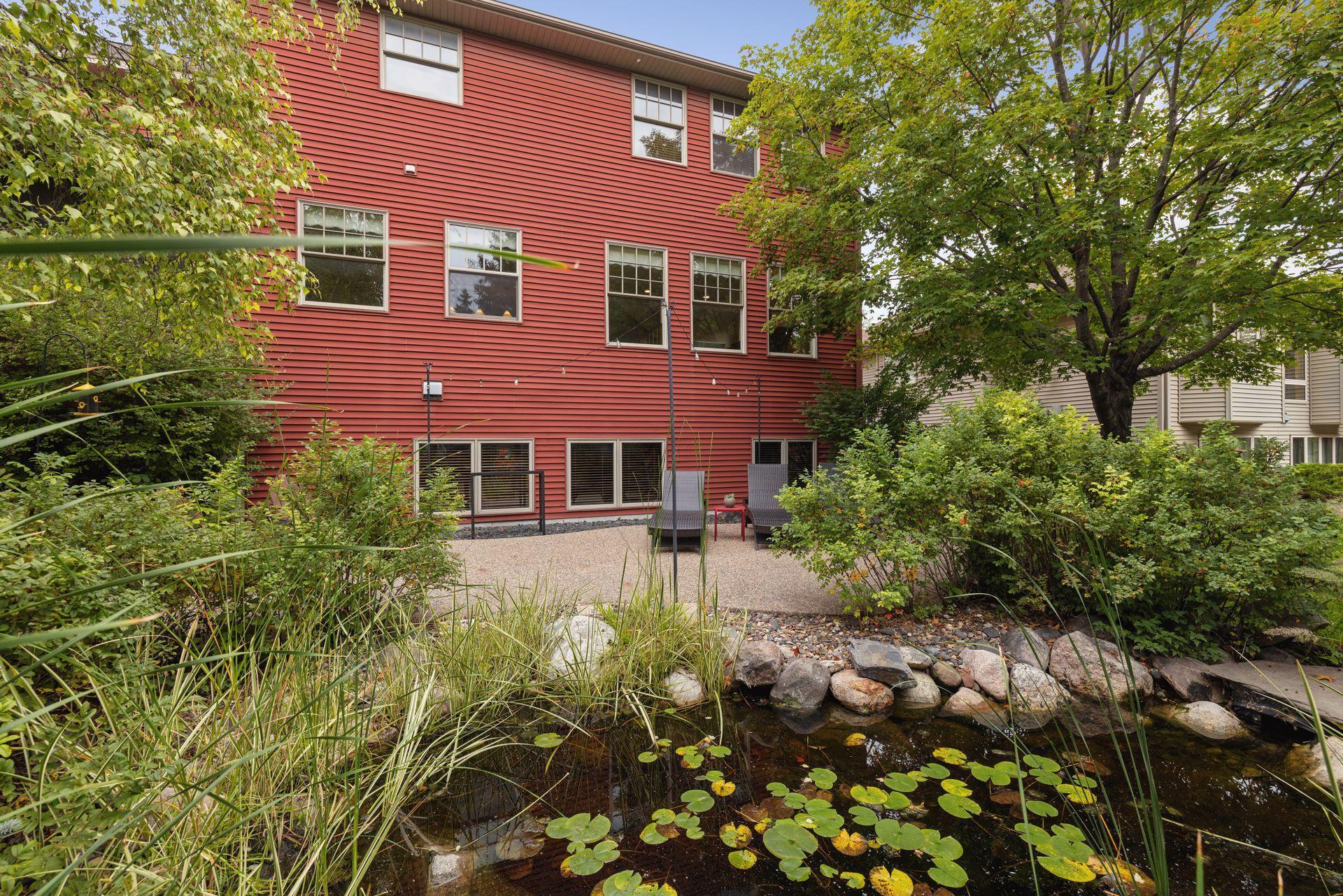Red-sided home with a patio and chairs beside a landscaped pond.