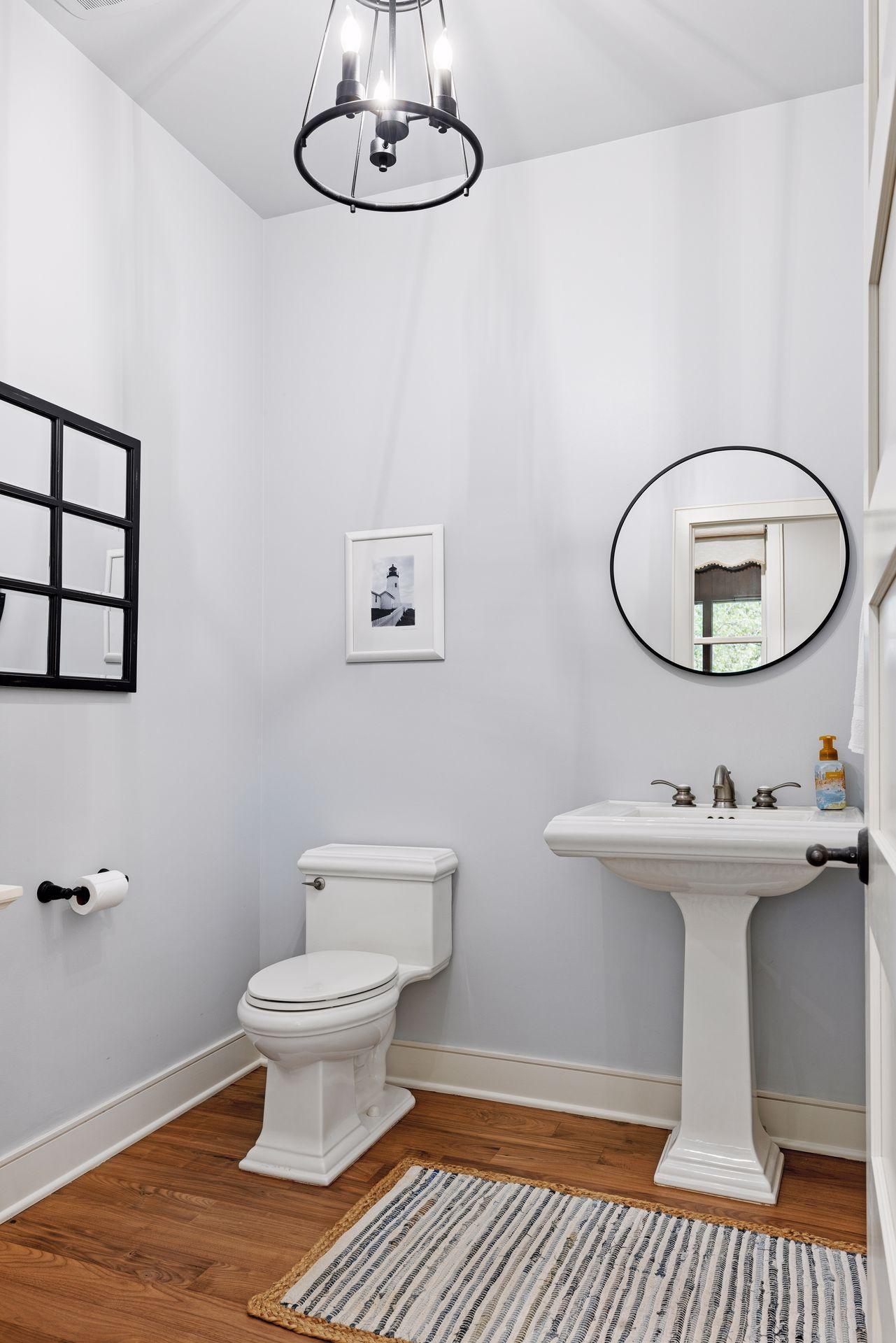 Half bathroom with a pedestal sink, round mirror, white toilet, and striped rug.