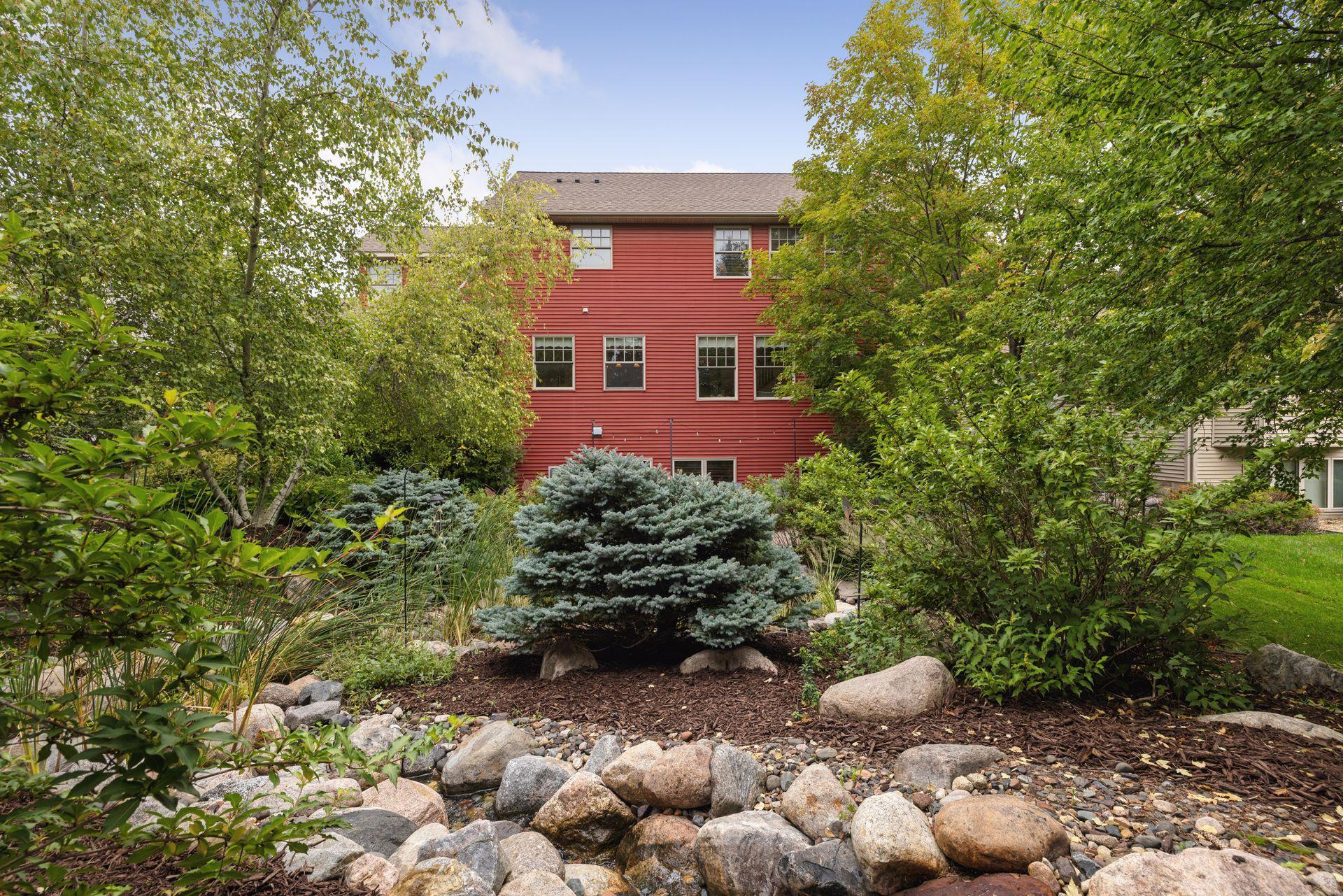 Red-sided house behind mature trees, with a blue spruce and landscaped rock garden in the foreground.