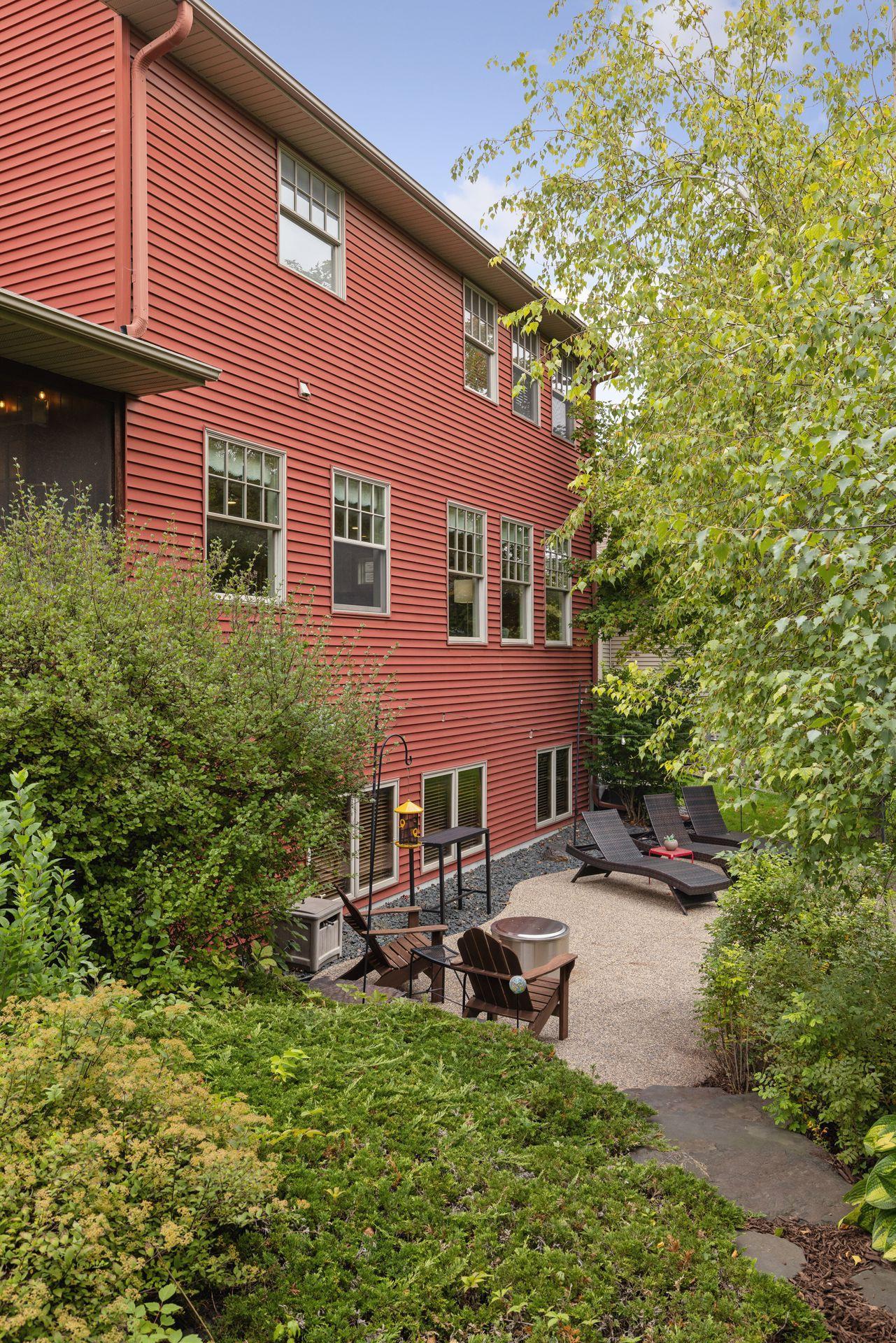 Red-sided home with a landscaped patio, lounge chairs, and leafy trees.